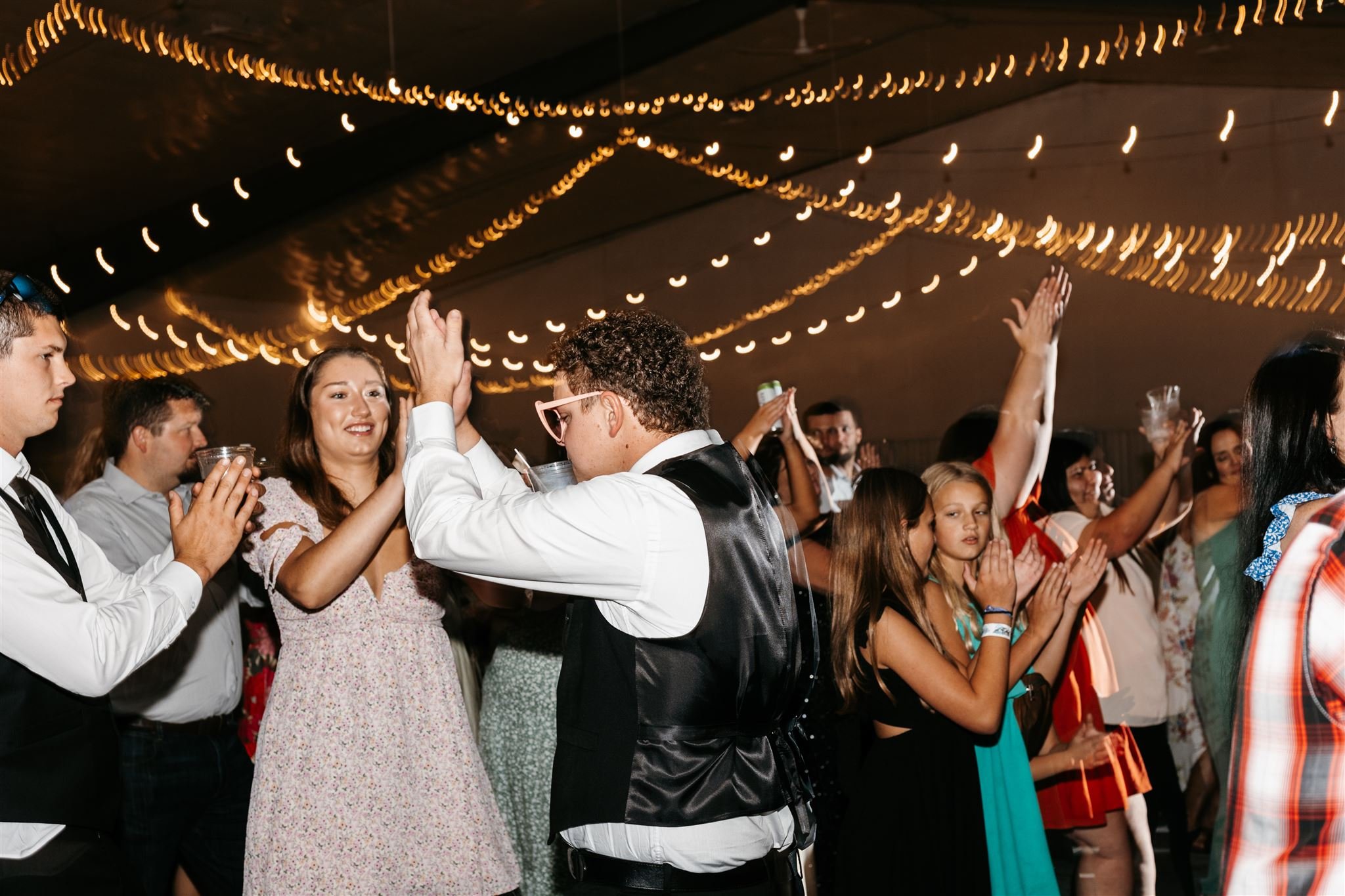 People dancing and celebrating at a party under hanging string lights, enjoying drinks and socializing.
