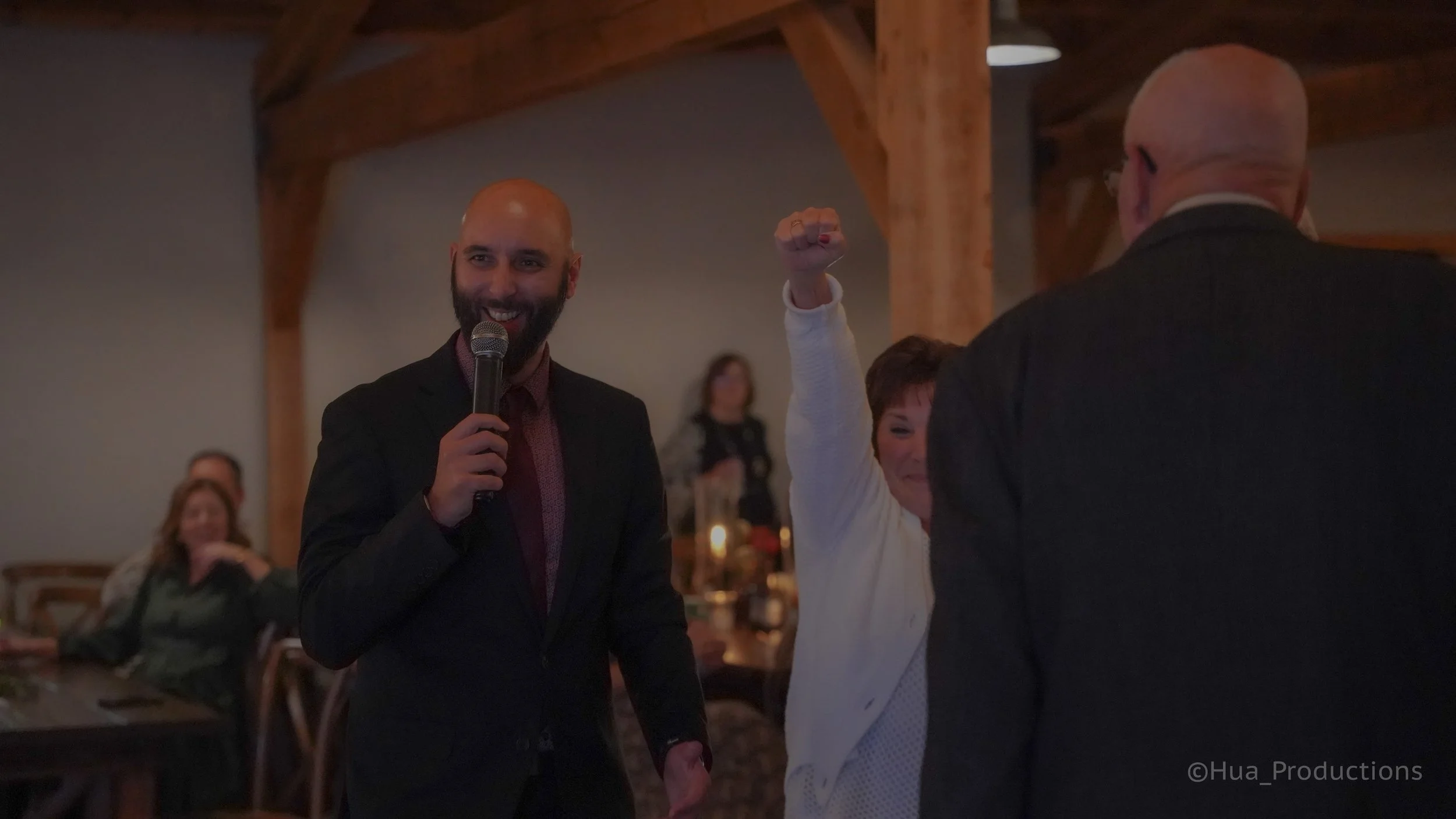 A man with a beard in a suit holding a microphone, smiling at a gathering with people in the background, including a woman raising her fist in the air.