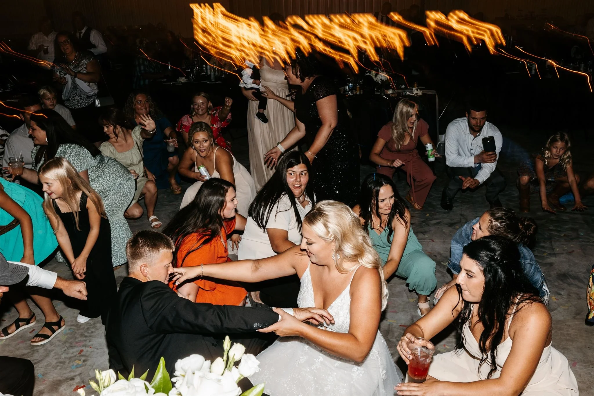 A wedding reception with a bride and groom dancing, surrounded by guests sitting on the floor and enjoying the celebration. Some guests are taking selfies and others are dancing, with bright orange and yellow light trails in the background.
