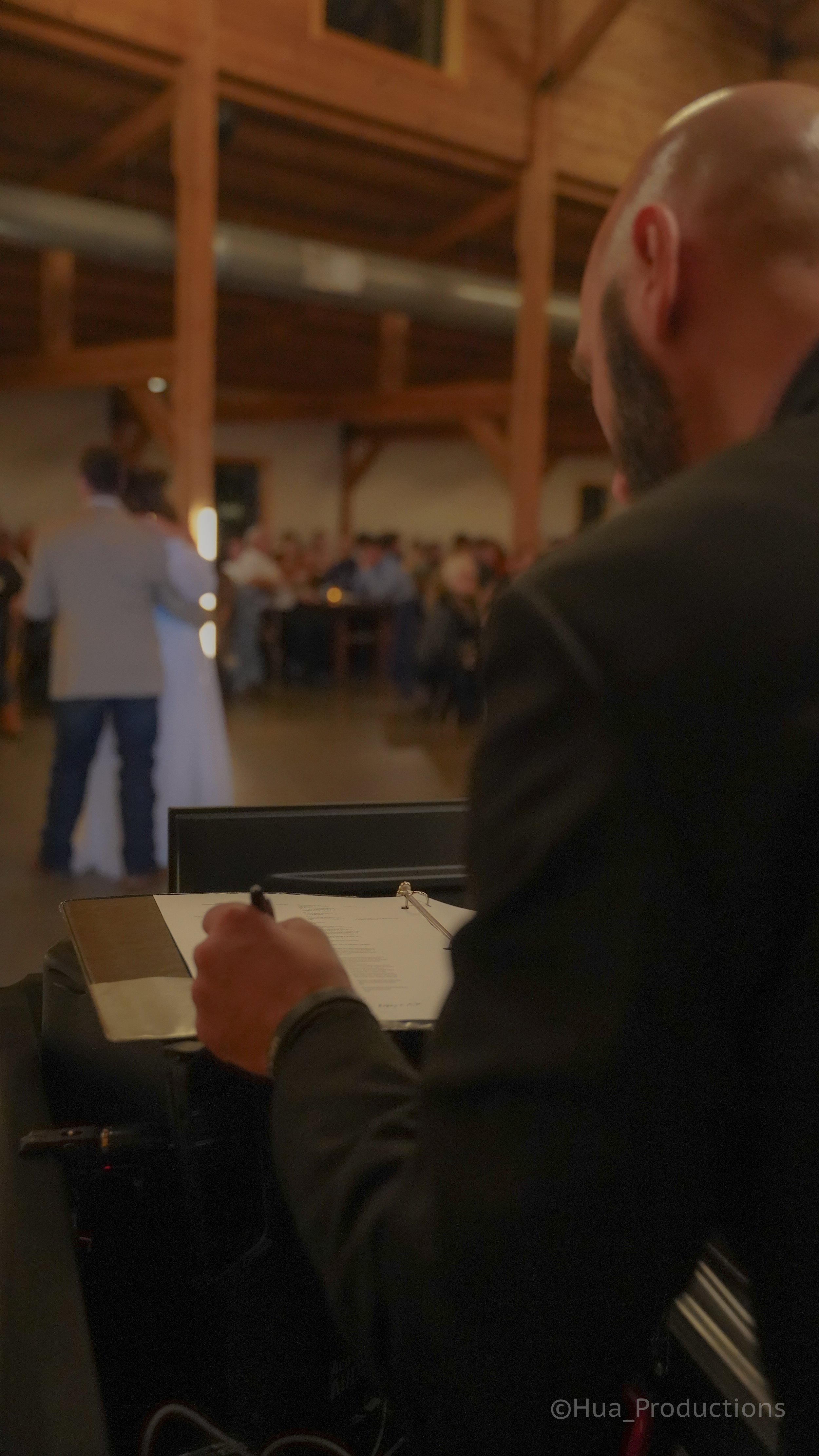 A man with a cigar in his mouth and a black suit sitting at a table with a notebook, overlooking a wedding reception in a rustic hall with wooden beams, where a couple is dancing.