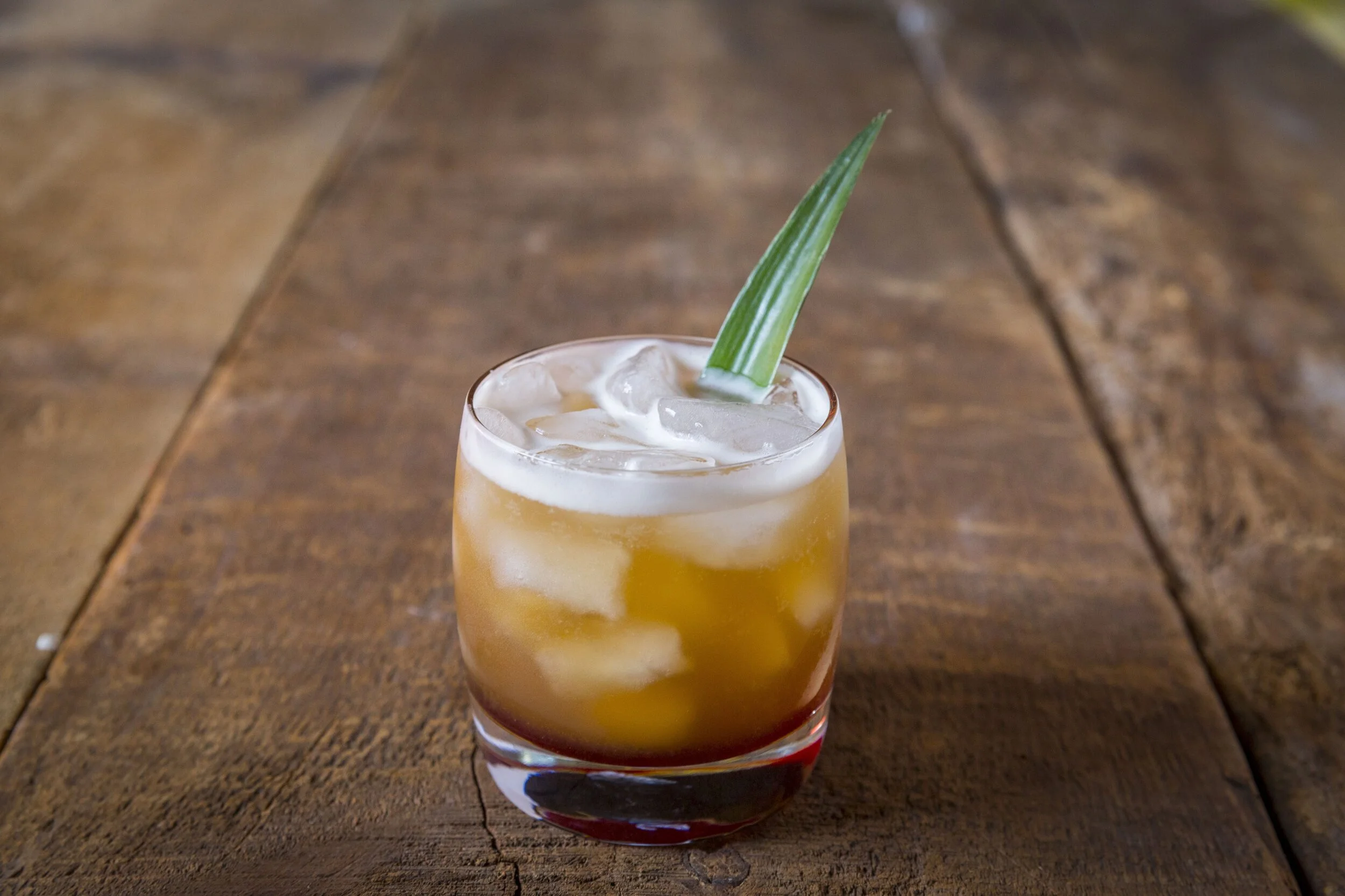 A cocktail in a short glass with ice, garnished with a green leaf, placed on a wooden table.