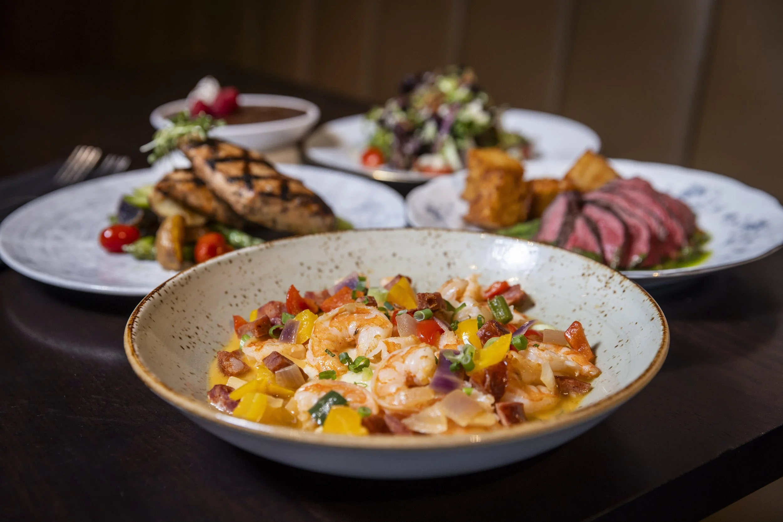 A variety of plated dishes including shrimp with vegetables in the foreground, grilled chicken with cherry tomatoes and greens, roasted sweet potatoes, sliced steak, and a salad in the background.