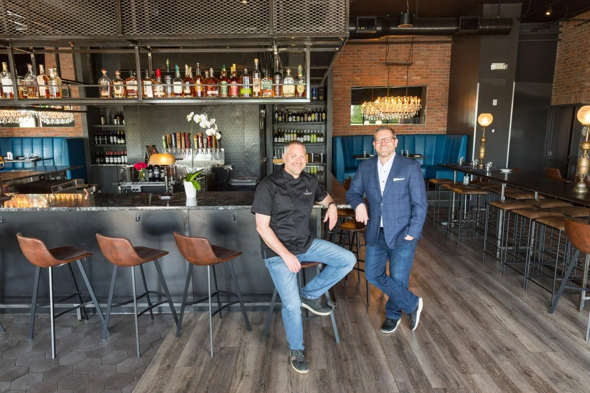 Two men in a restaurant, one in a chef's coat and the other in a suit, standing by a bar counter with stools. The restaurant has a modern design with brick walls, wine bottles on shelves, and elegant lighting.