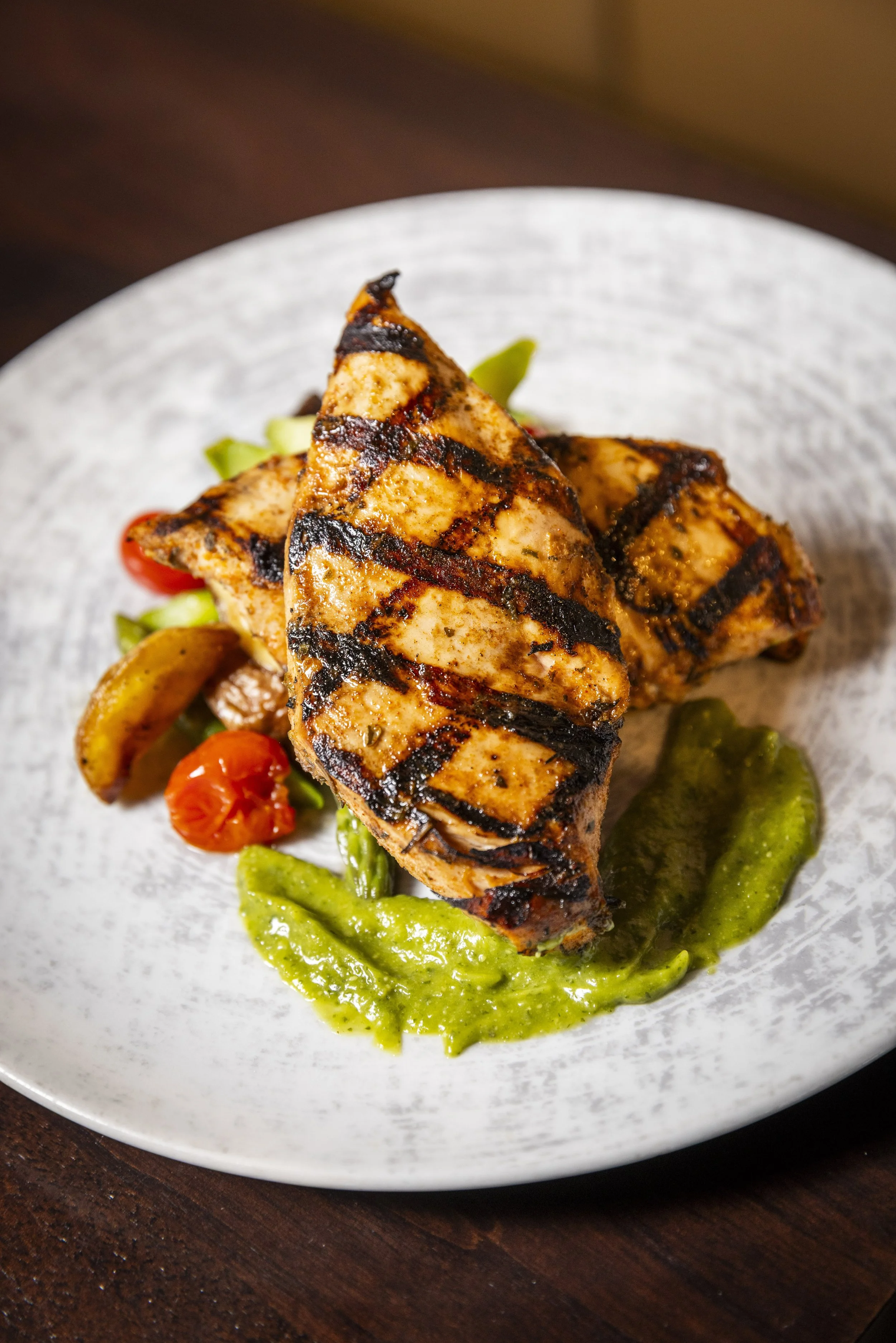 Grilled chicken with char marks served with sautéed asparagus, charred tomatoes, basil butter sauce, and microgreens on a white plate.