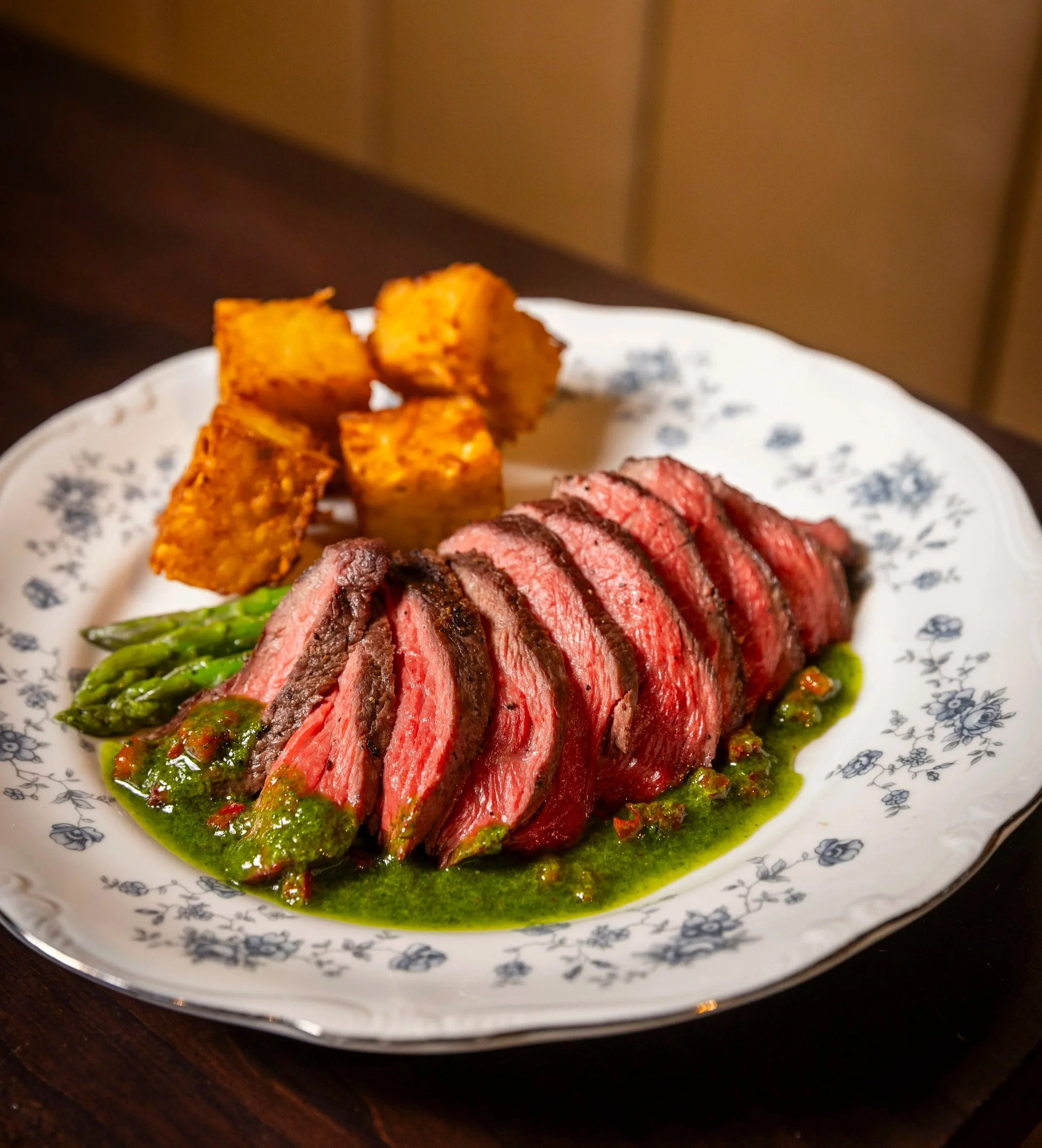 Slices of grilled sirloin steak with green chimichurri and crispy hash brown squares on a white floral plate.