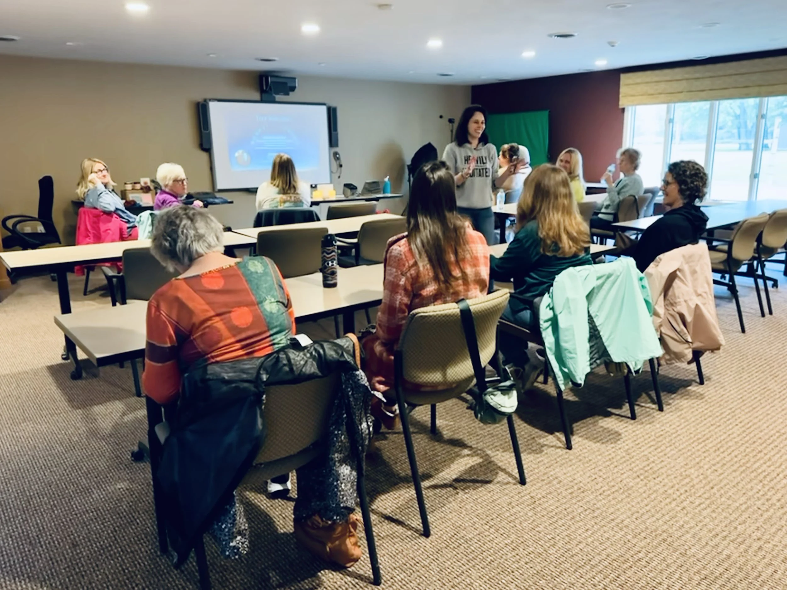 Women attending a workshop or class in a conference room, with a woman standing and speaking at the front near a large screen.