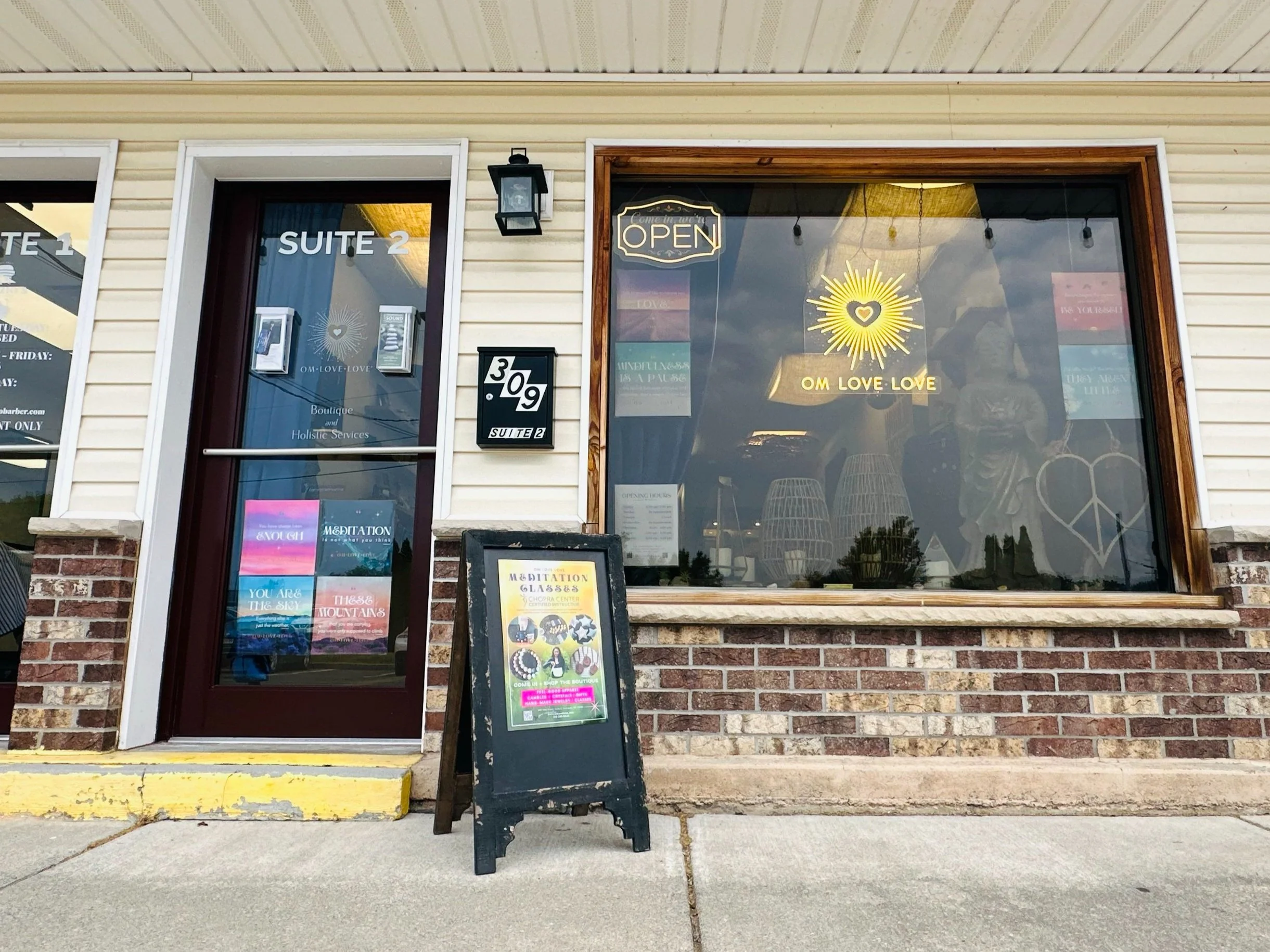 The exterior of a shop with a large front window and a door labeled 'SUITE 2'. A black sign with '309 SUITE 2' is on the wall, and there's an easel sign outside advertising meditation classes. The window has a yellow sign with 'OM LOVE LOVE' and a heart with rays of light. An 'OPEN' sign is hanging above. The building has cream-colored siding and brick at the bottom.