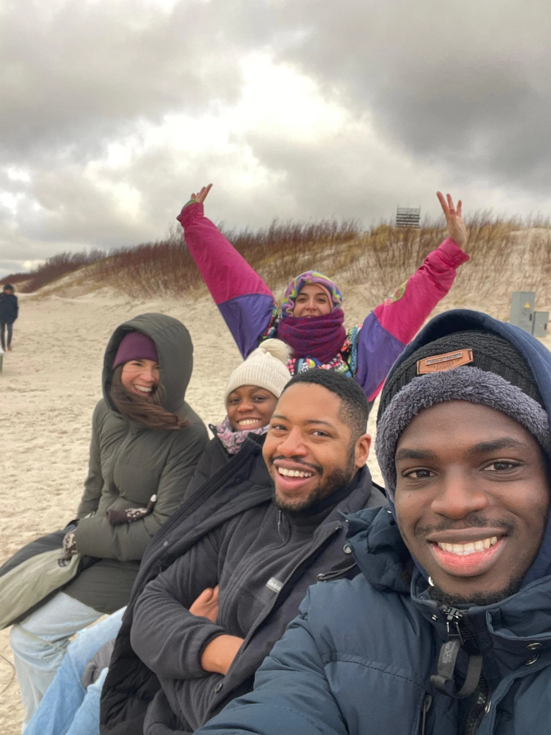 A group of five friends taking a selfie on a sandy beach with dunes and cloudy sky in the background. They are smiling, dressed in warm clothing.
