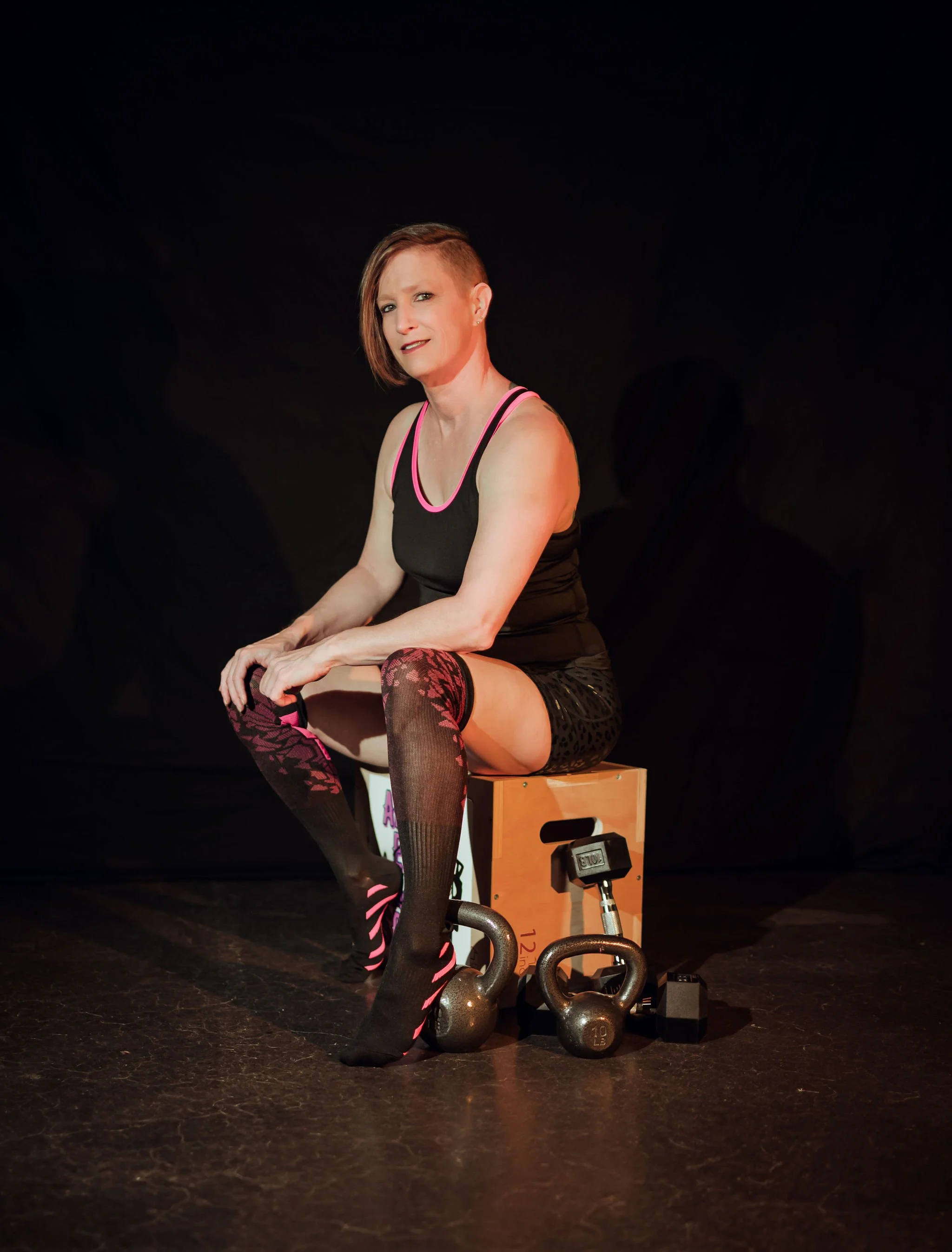 A woman in workout attire sitting on a wooden box, surrounded by kettlebells and a dumbbell, against a black background.