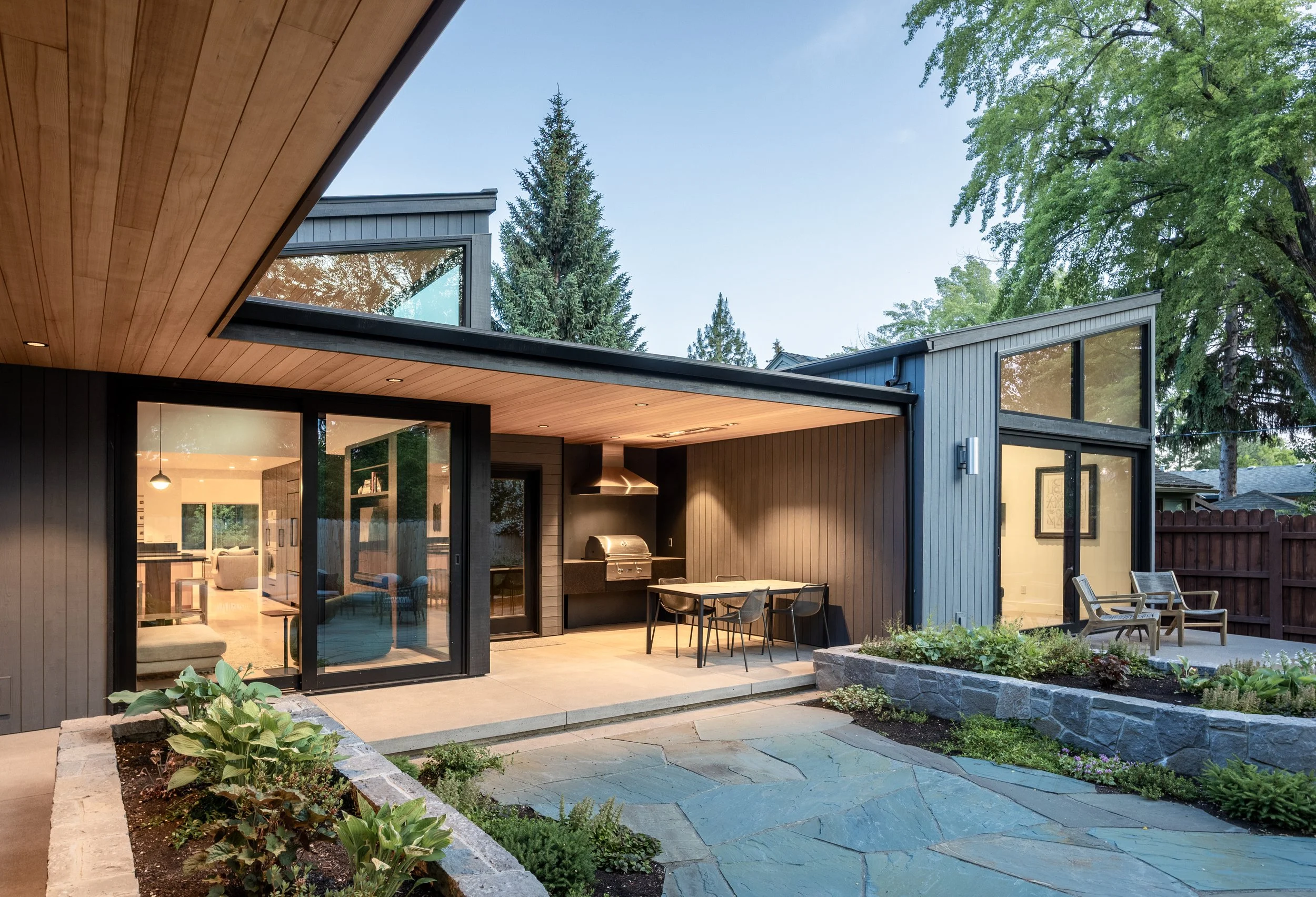 Modern house exterior with a patio, outdoor dining area, built-in grill, garden beds, and a backyard with trees. In downtown Bend Oregon
