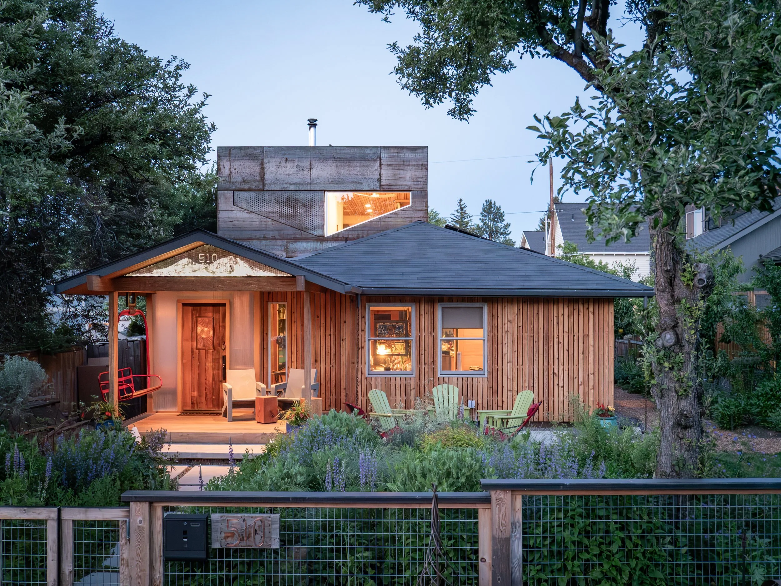 A cozy wooden house with a small front porch, sitting in a lush garden with outdoor furniture, large mature trees, and neighboring houses in the background. In Bend Oregon