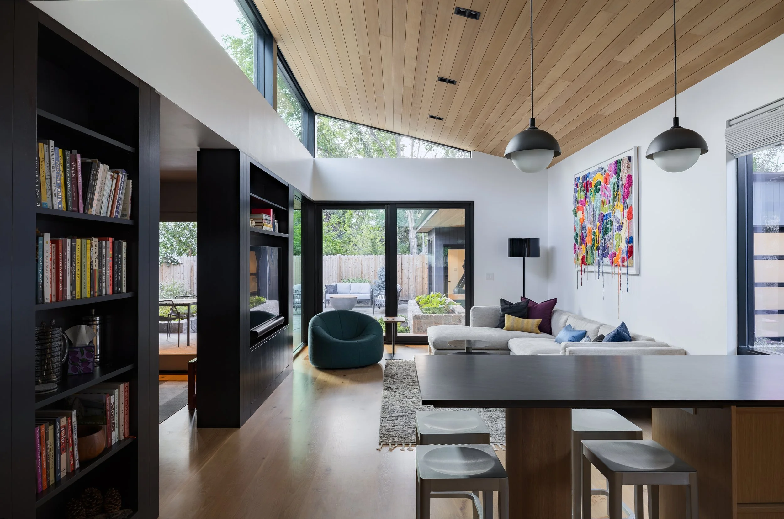Modern living room with wooden ceiling, large windows, white walls, a sectional sofa with colorful pillows, a black bookshelf, a television, two hanging black and white pendant lights, and an outdoor patio visible through sliding glass doors. In Bend