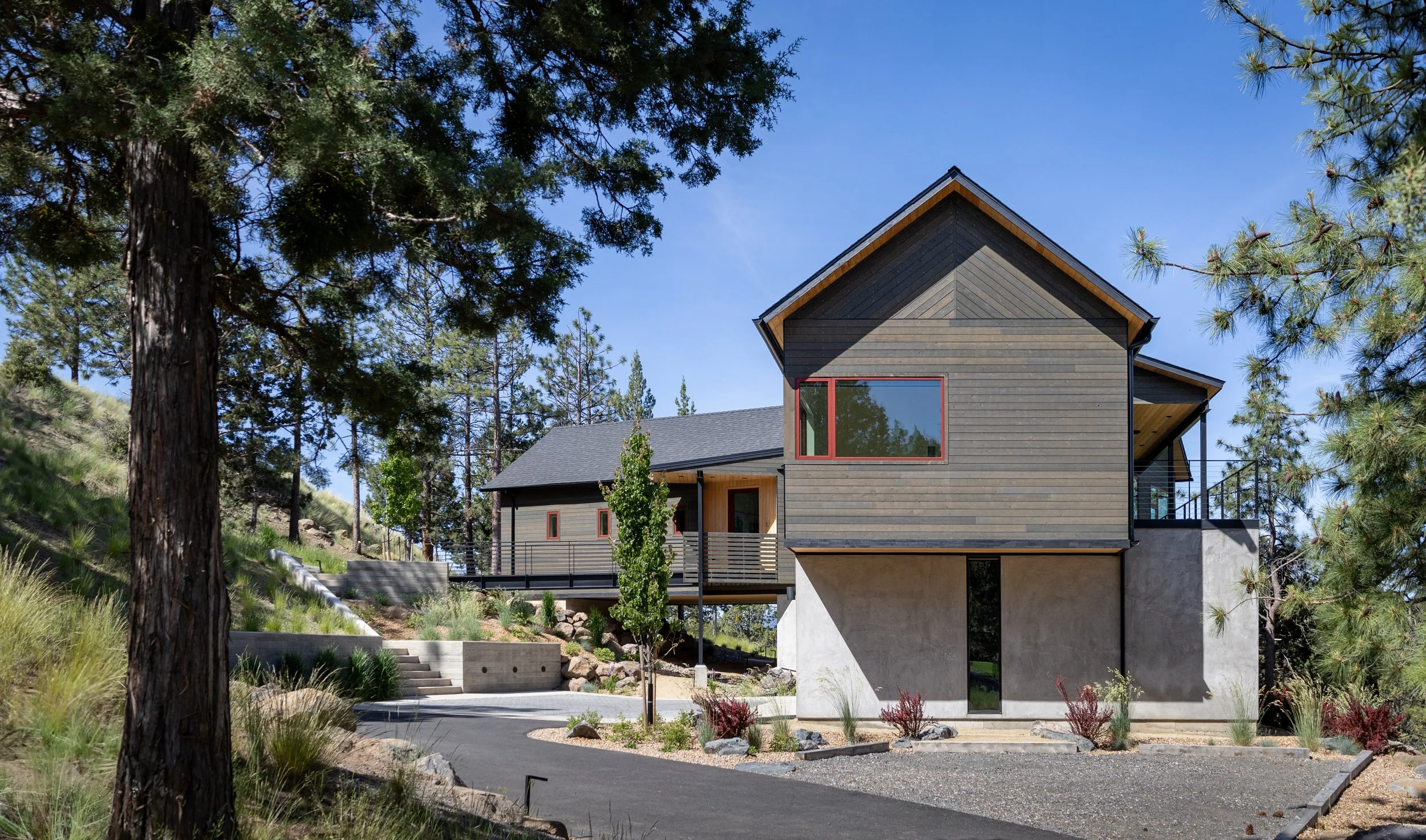 Modern house with wooden siding, concrete foundation, large windows, surrounded by trees and landscaping, on a sunny day. In Bend Oregon