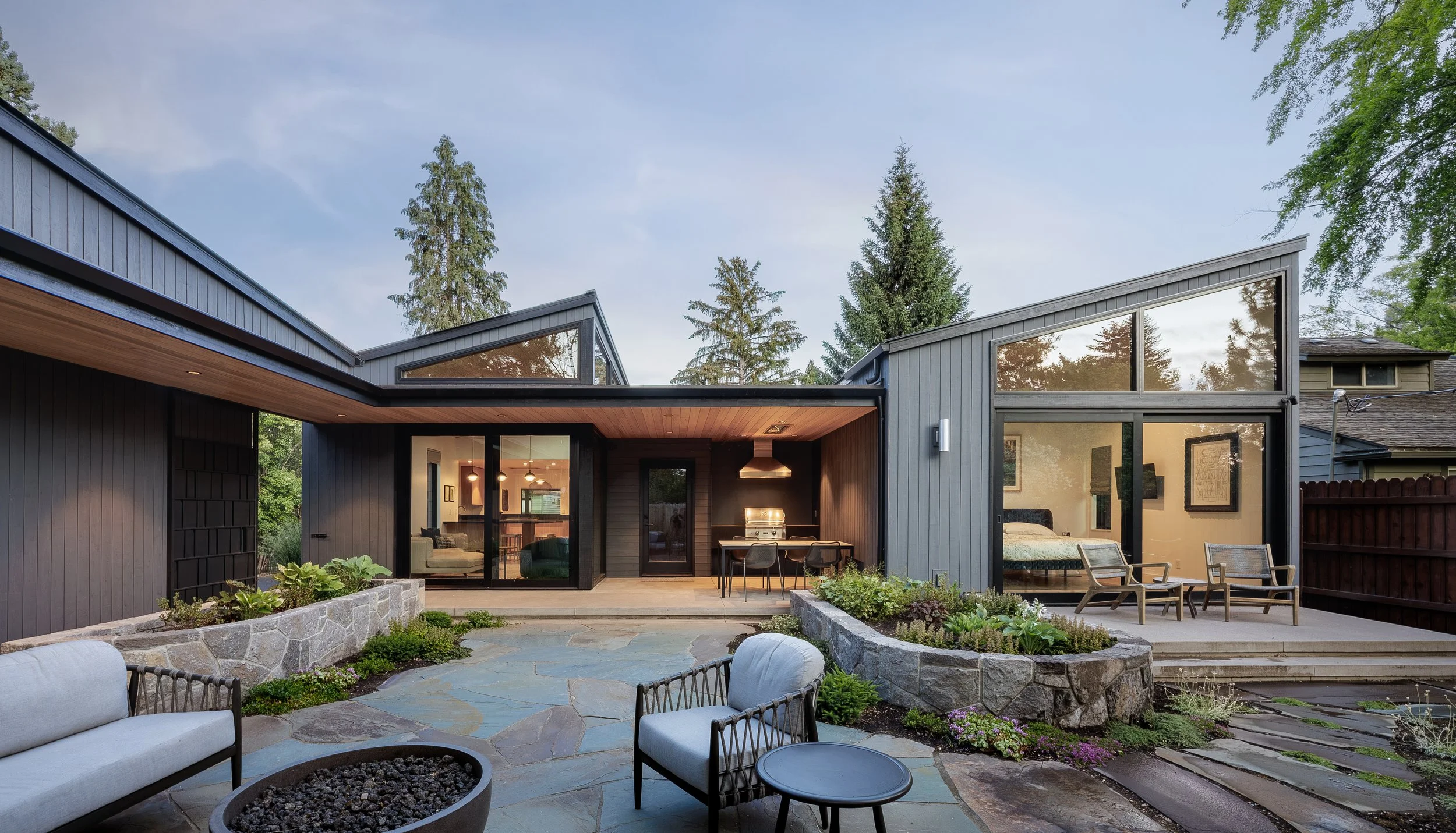 Modern house with large glass windows, outdoor patio with seating, garden beds, and trees in the background. In Bend Oregon