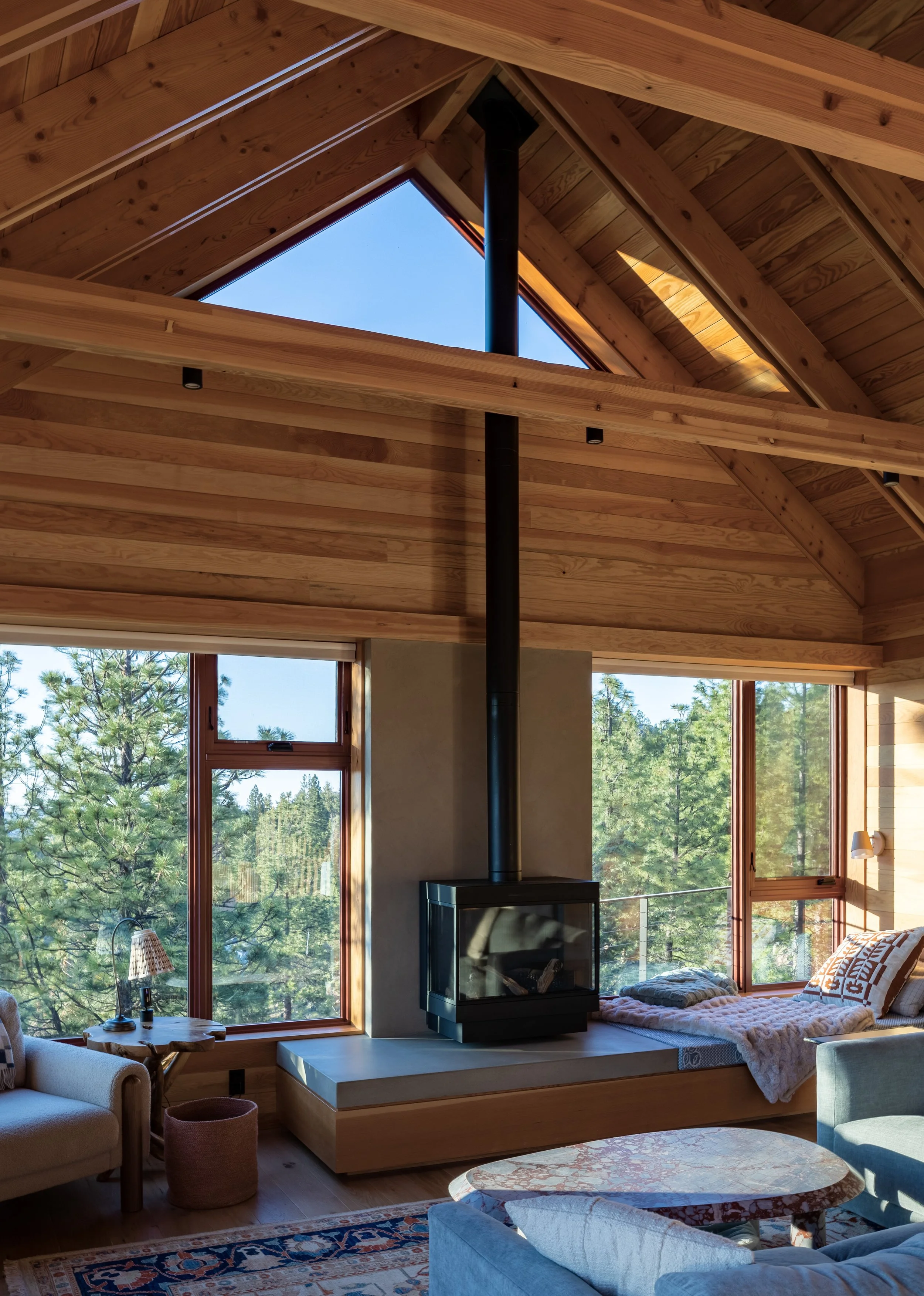 Living room with wooden vaulted ceiling and large windows showing trees outside. A black wood stove is in the center, with a built-in corner bench, a sofa, and a rug in the room. In Bend Oregon