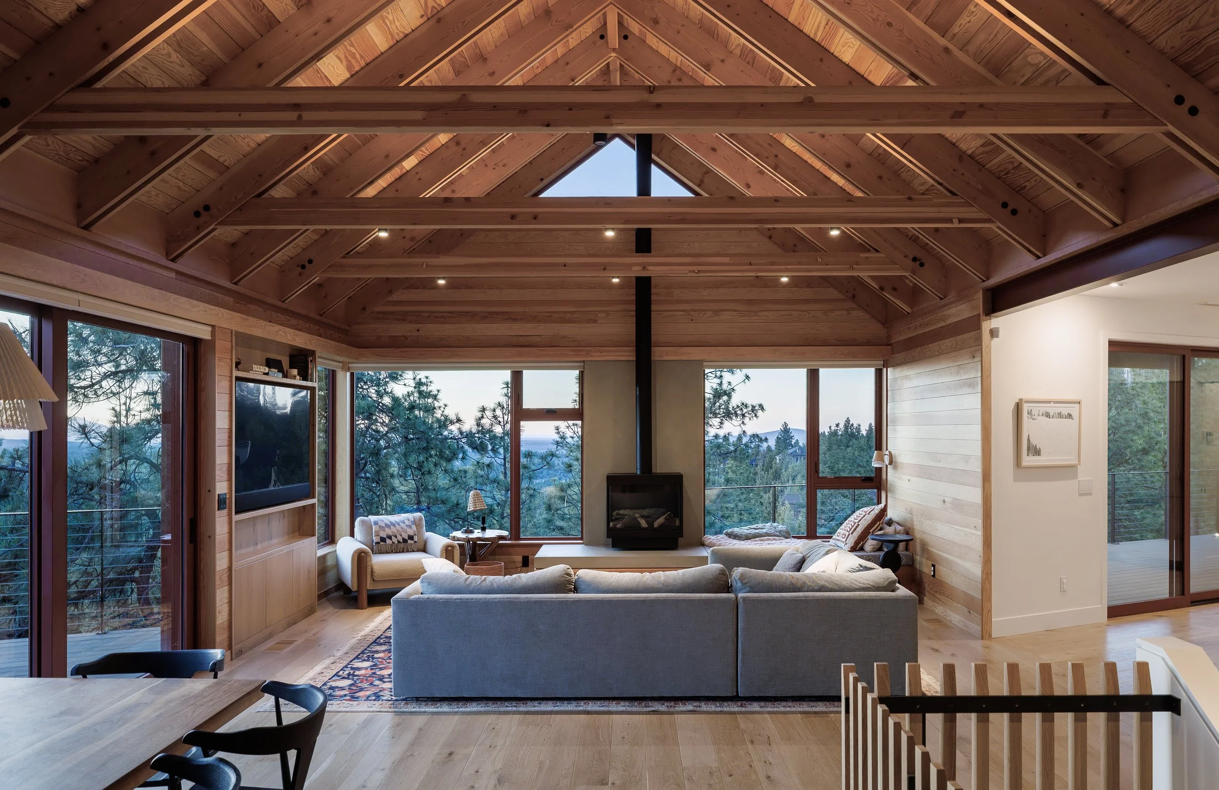 Living room with wooden vaulted ceiling, large windows, and a fireplace. Contains a gray sofa, armchair, and wooden furniture. In Bend Oregon
