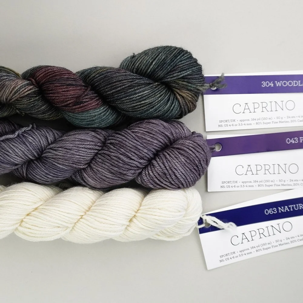 Three skeins of Caprino yarn in Woodland (dark multicolor), Black Plum (purple), and Natural (off-white) shades are arranged side by side with labeled tags visible.