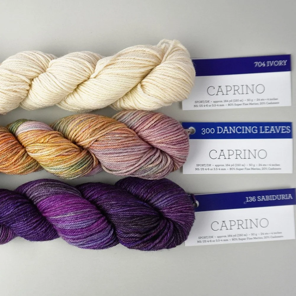 Three skeins of Caprino yarn in ivory, pink, and purple shades are stacked on a light surface with labeled tags attached.