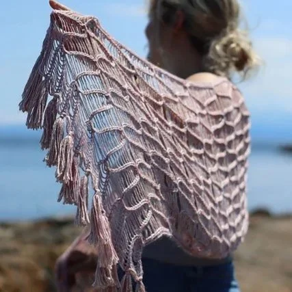  Person holding a pink crocheted shawl with tassels over their shoulders, standing by the sea. 
