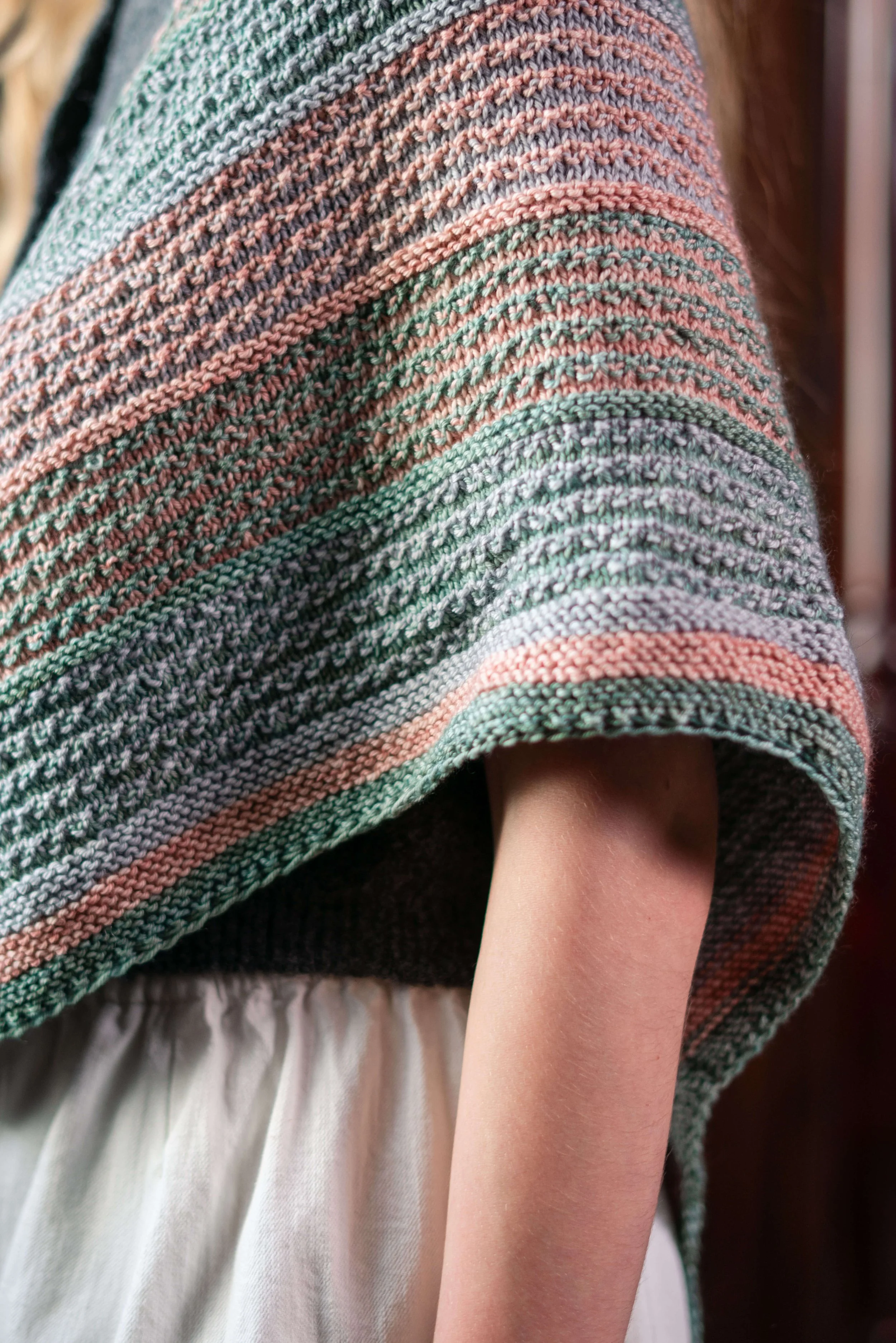 A knitted shawl with horizontal stripes of green, pink, and grey yarn is draped over a person's shoulders. The person is wearing a white dress.