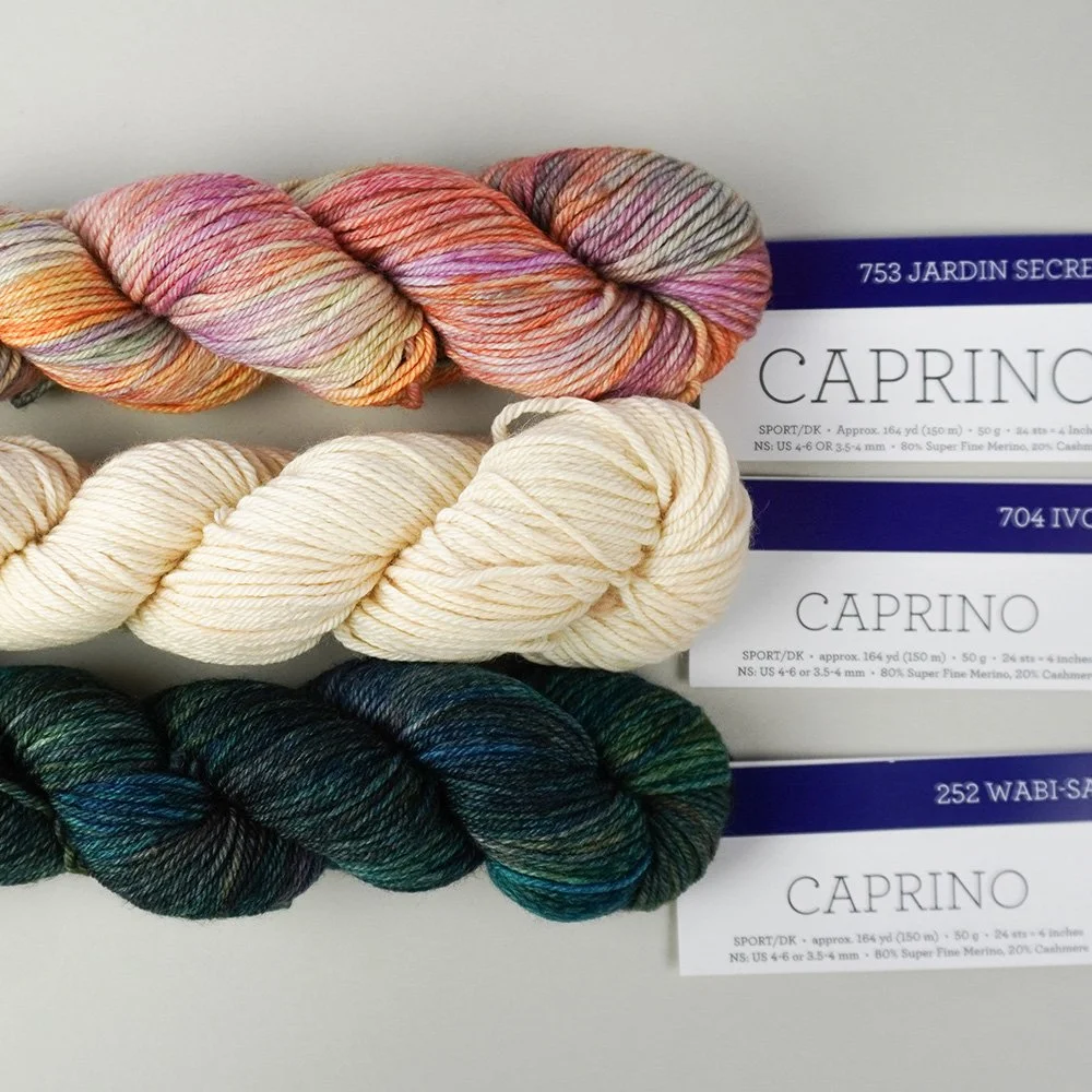 Three skeins of Caprino yarn in ivory, pink, and green shades are stacked on a light surface with labeled tags attached.