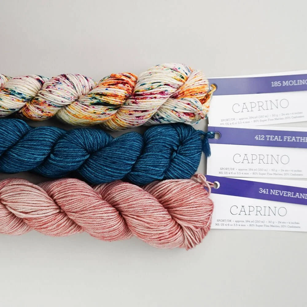 Three skeins of Caprino yarn in variegated colors, blue, and pink shades are stacked on a light surface with labeled tags attached.