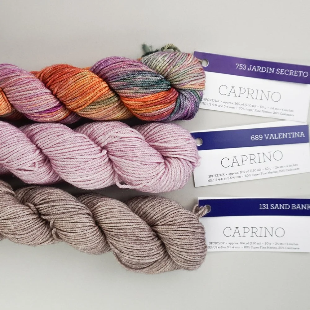 Three skeins of Caprino yarn in variegated pink and orange, Pink, and sand bank shades are stacked on a light surface with labeled tags attached.