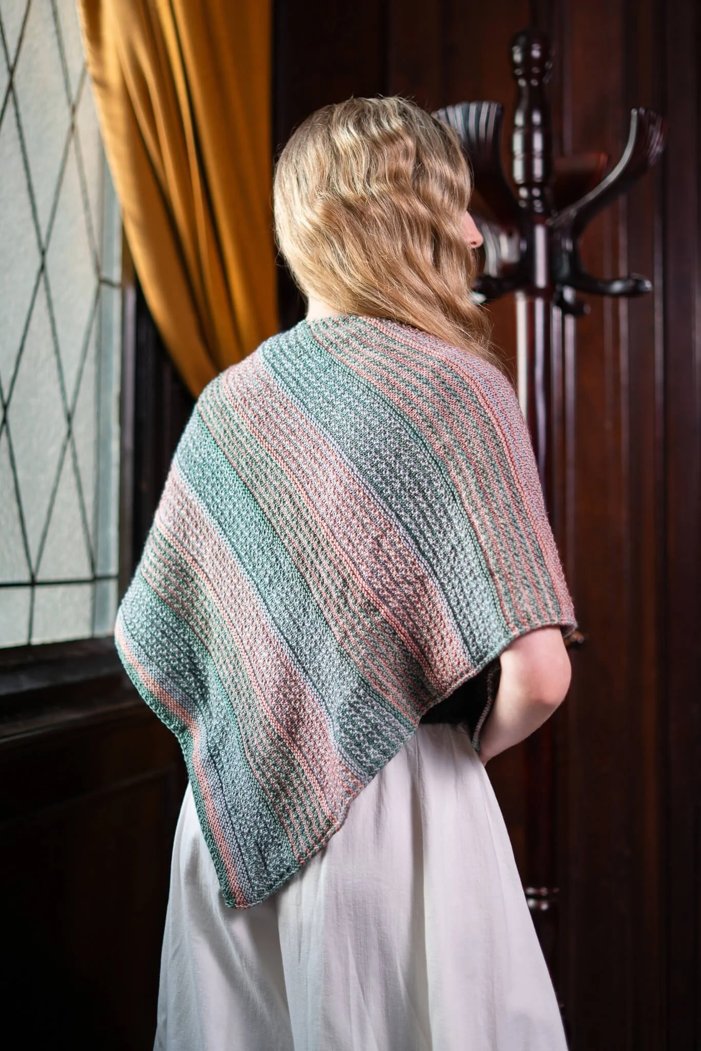 A person with wavy blonde hair wearing a striped knit shawl and a white dress stands indoors, facing away from the camera near wood paneling and a window with yellow curtains.