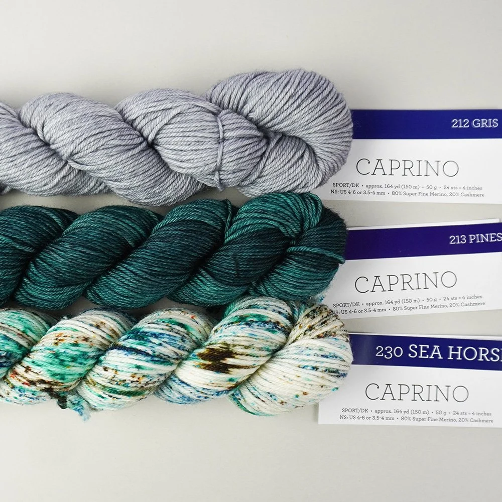 Three skeins of Caprino yarn in variegated green, gray, and green shades are stacked on a light surface with labeled tags attached.