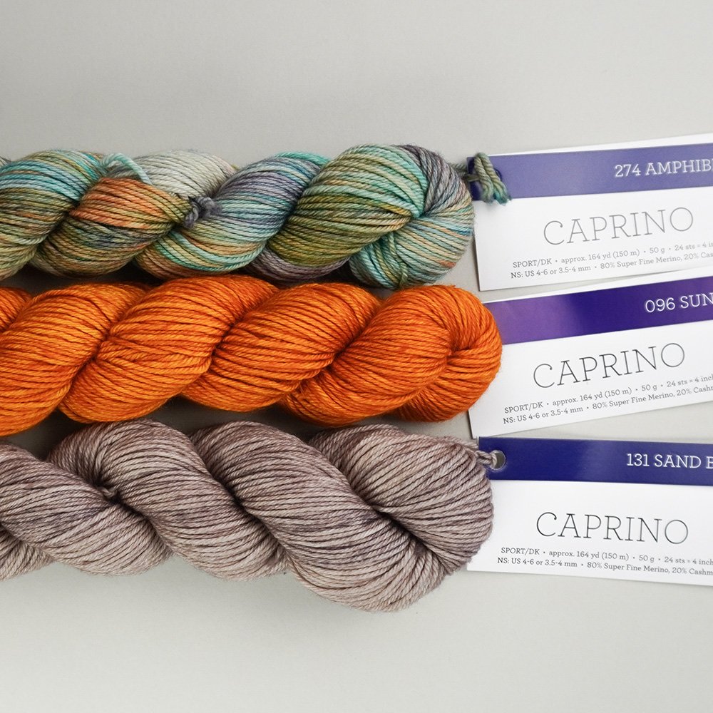 Three skeins of yarn in multicolor, orange, and taupe shades are labeled as Caprino in colors 274 Amphibian, 096 Sunset, and 131 Sand Bank, laid side by side on a gray surface.