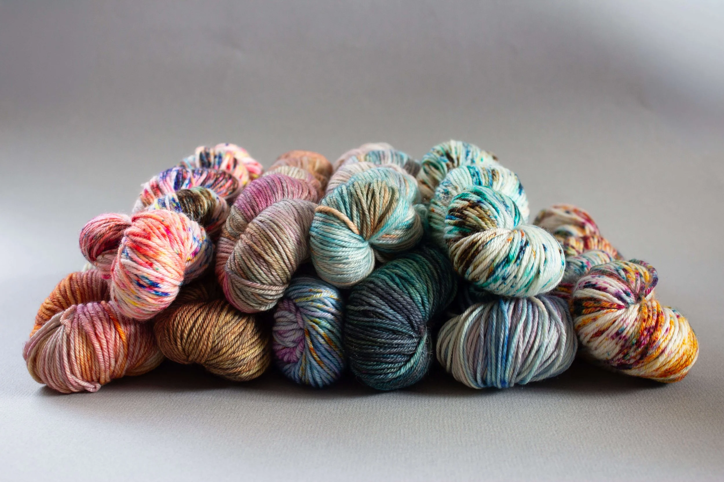 A pile of hand-dyed yarn skeins in various colors including pink, orange, teal, green, and cream, against a neutral gray background.