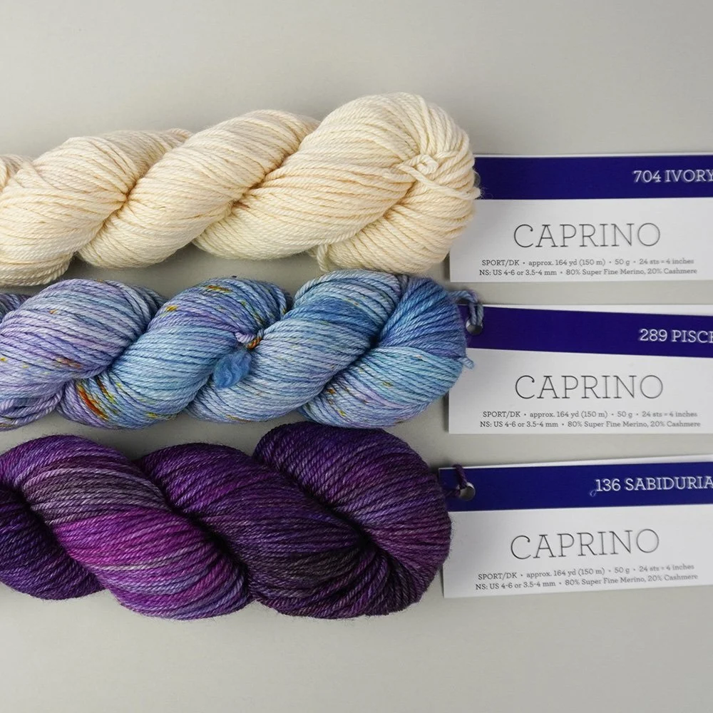 Three skeins of Caprino yarn in ivory, blue, and purple shades are stacked on a light surface with labeled tags attached.