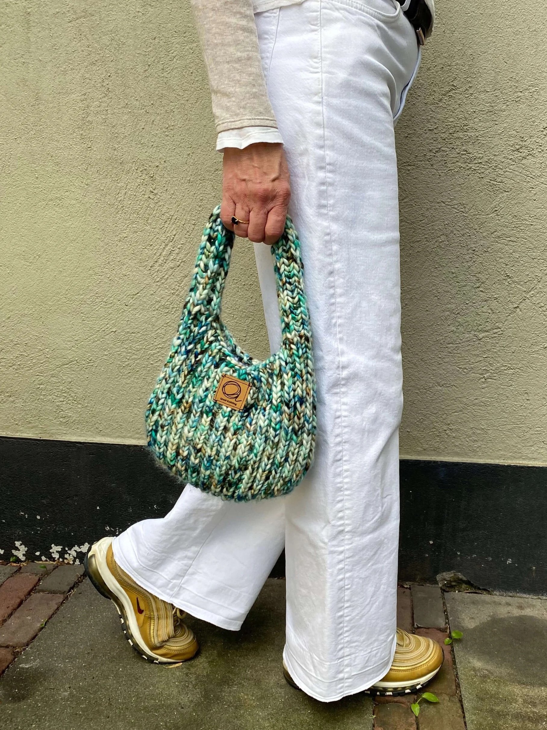 Person wearing white pants and a beige sweater holding one knitted handbag in green and multicolor yarn, Noventa from Malabrigo