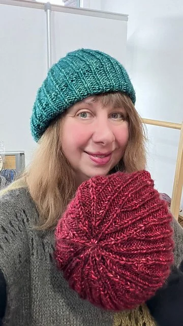 A person wearing a teal knitted hat and holding a red knitted hat.