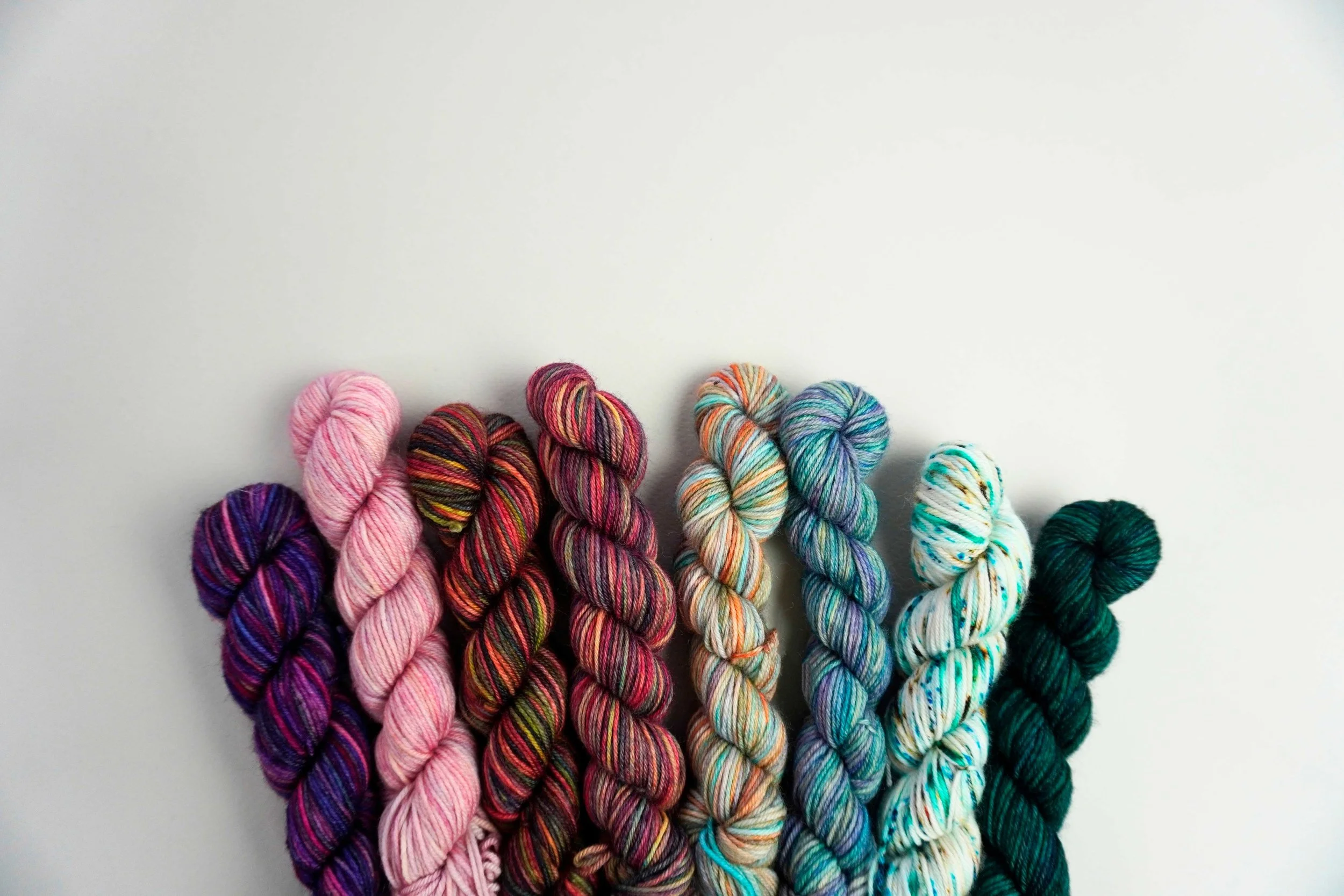 A row of eight skeins of variegated yarn in shades of purple, pink, red, orange, green, blue, teal, and dark green.