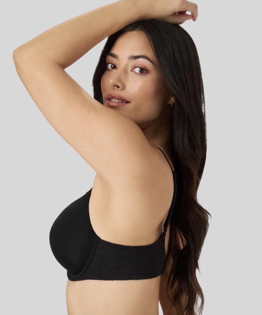 A woman with long dark hair wearing a black bra, posing with her right arm raised and resting on her head, looking at the camera against a plain gray background.