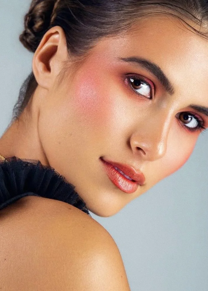 Close-up of a woman with styled dark hair, wearing pink and orange makeup, and a black ruffled garment, facing slightly to the right.