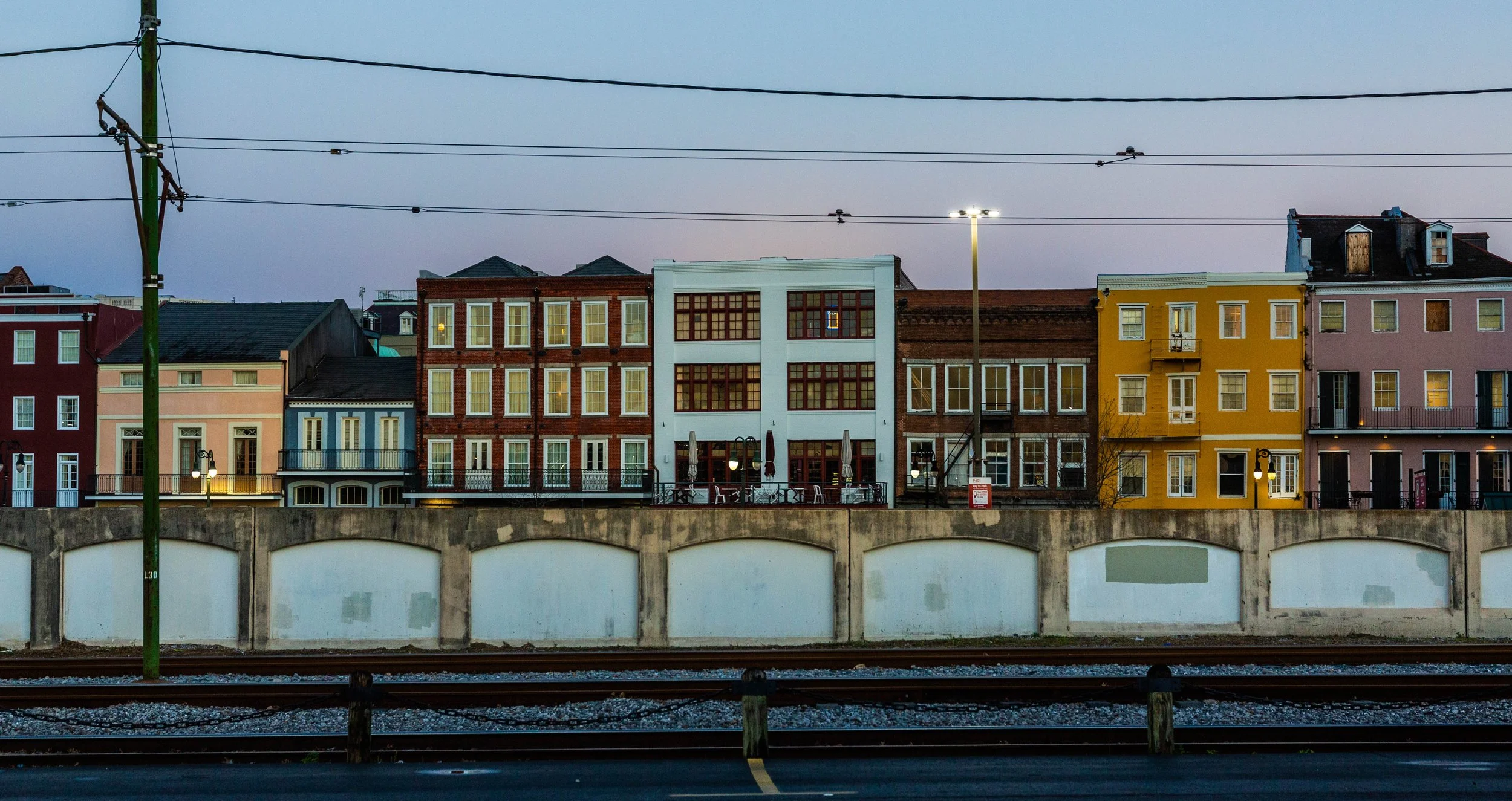 Colorful buildings on a city street viewed from across train tracks, with overhead power lines and a lamppost during dusk.