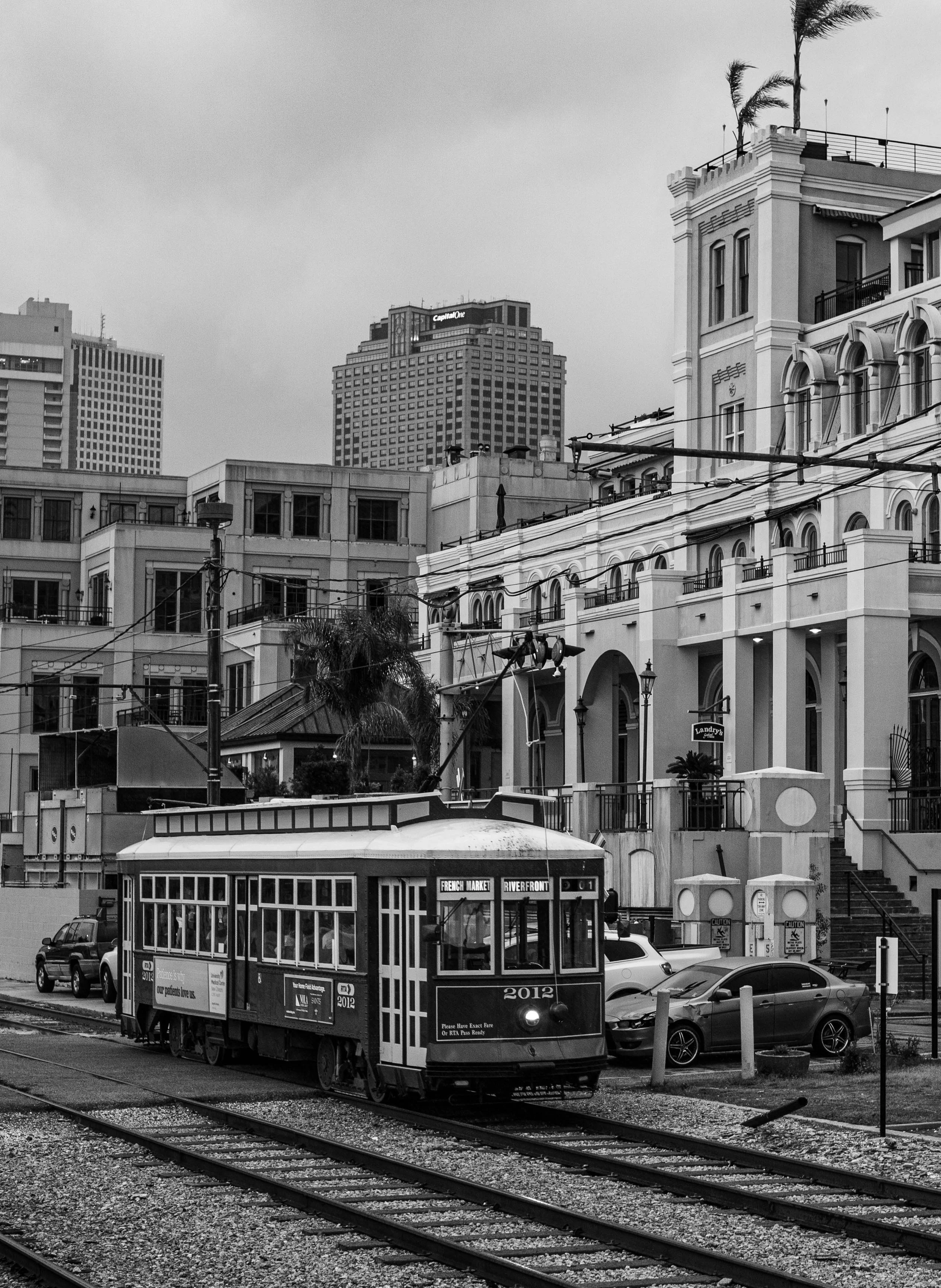 Black and white photo of a city street with a tram, cars, and tall buildings, including one with the Capital One logo, on a cloudy day.