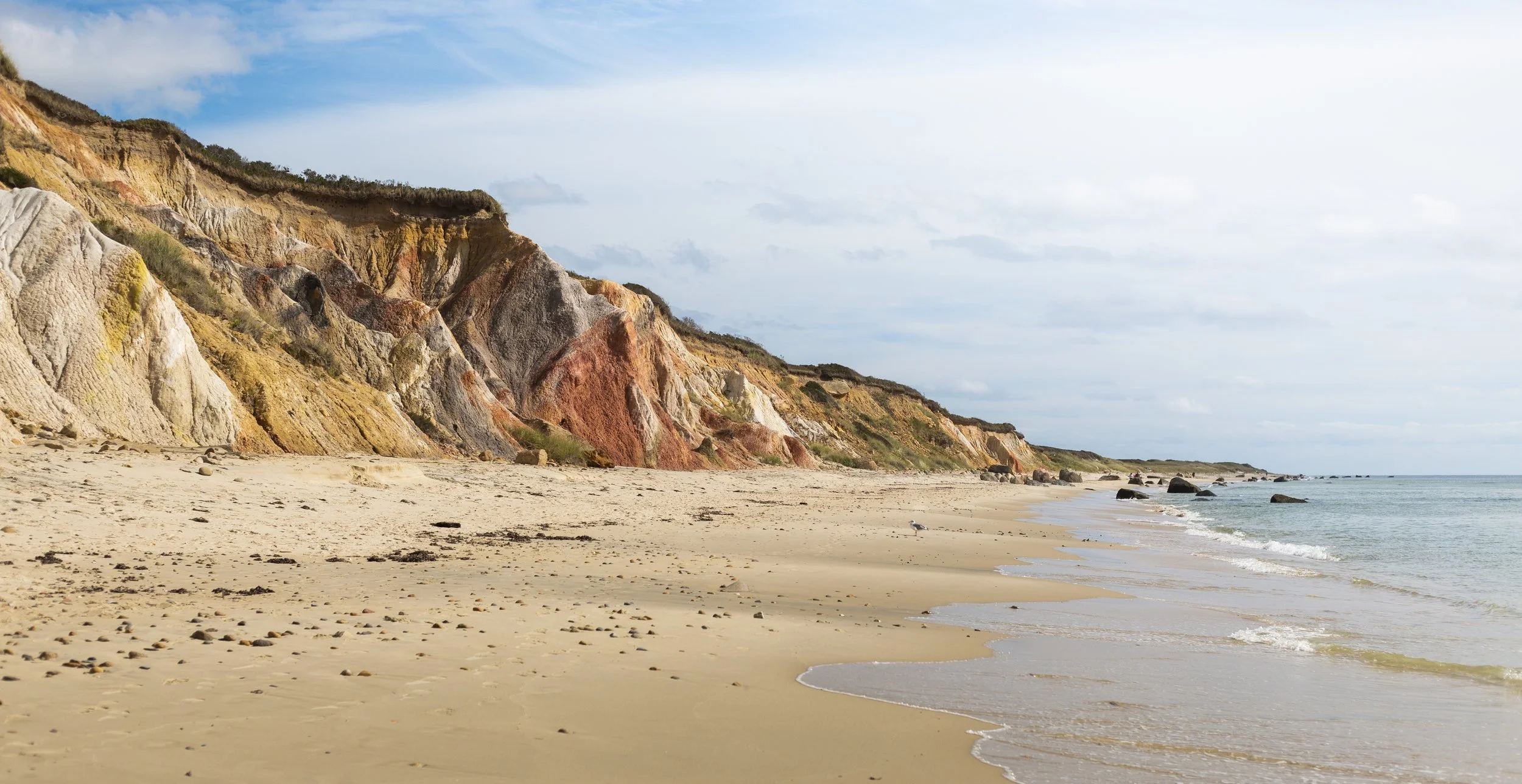 Scenic view of a sandy beach with colorful cliffs on the left and the ocean on the right, under a partly cloudy sky.