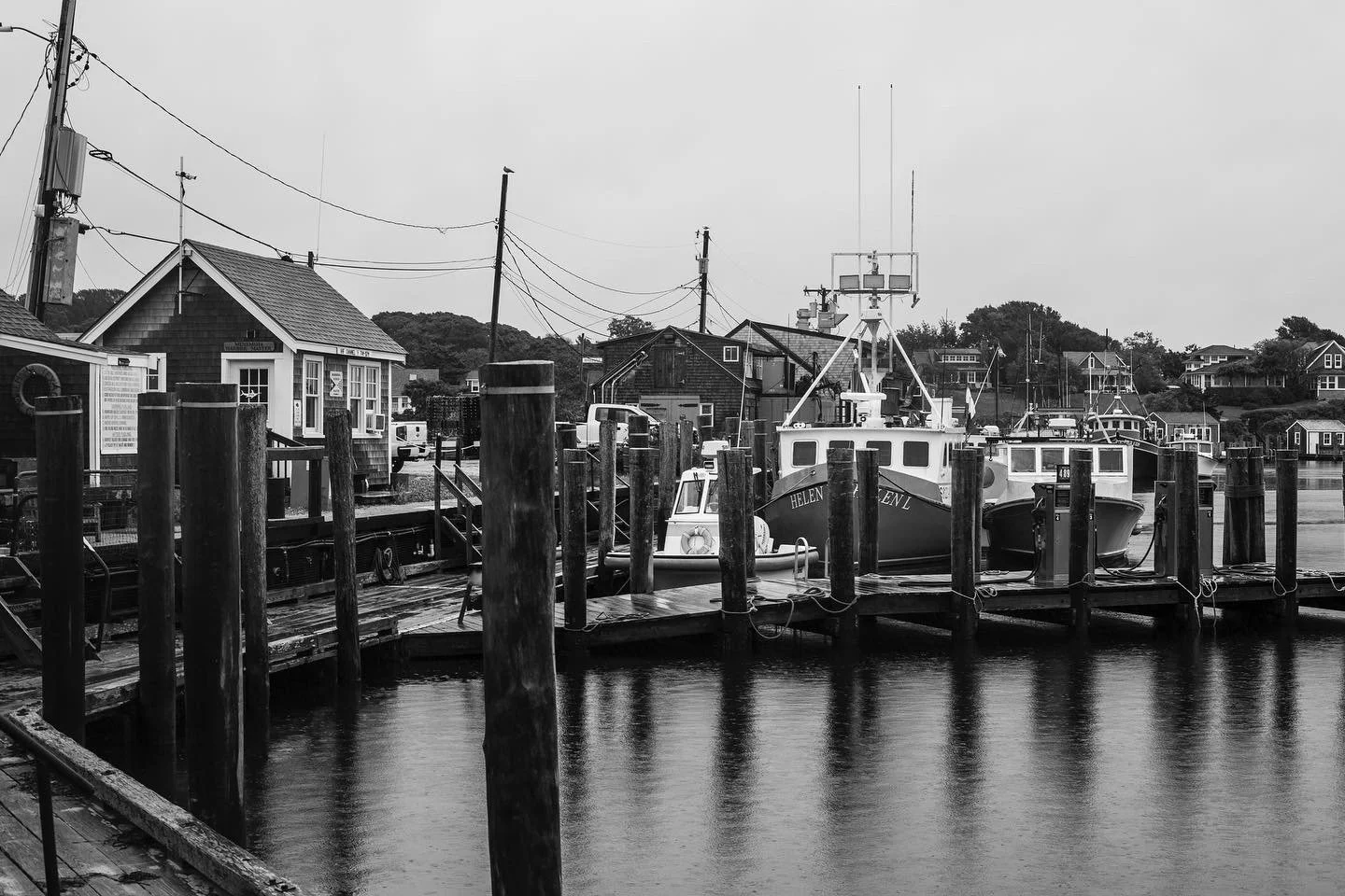 Black and white photo of boats docked at a small harbor with houses on a hill in the background.