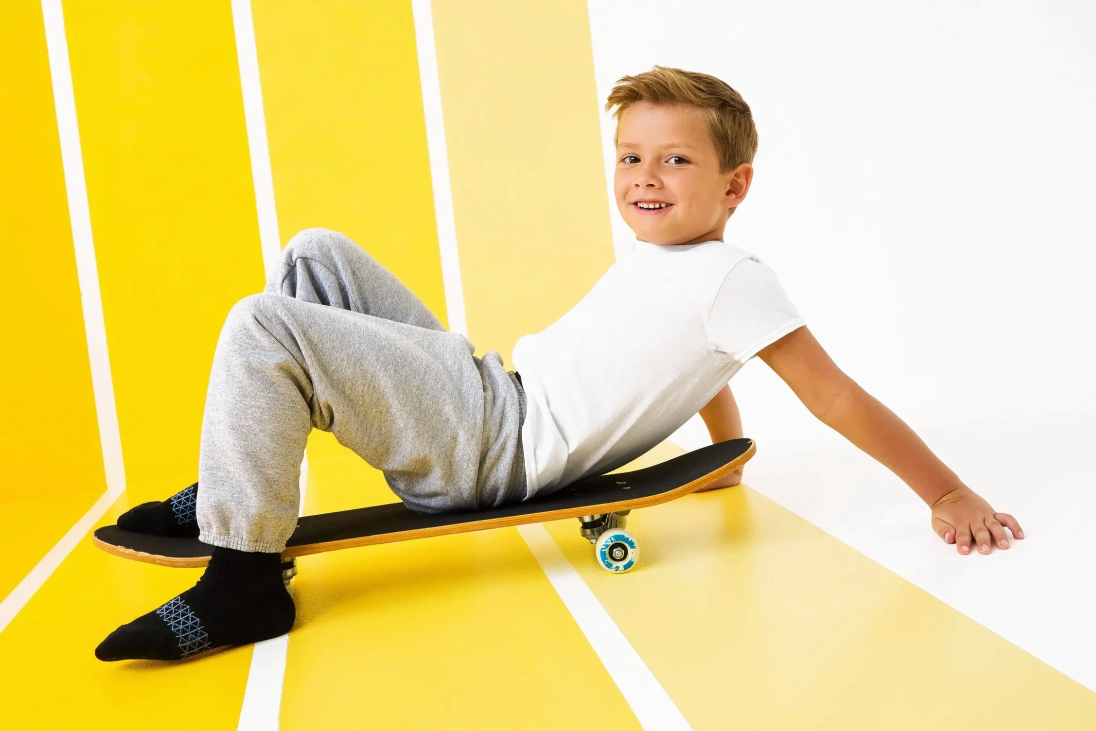 A young boy sitting on a skateboard with a bright yellow background and white lines, smiling at the camera.