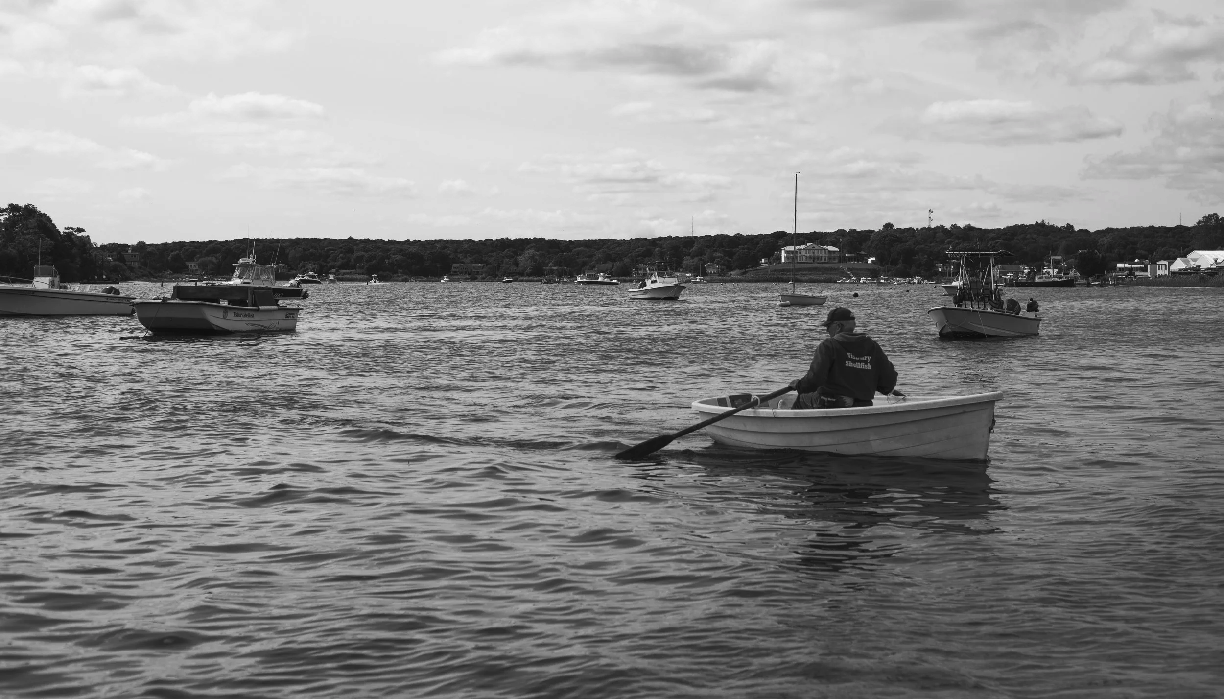 A person in a small rowboat on a river, wearing a jacket that says 'Treasure Shellfish', while several boats float on the water, with houses and trees on the riverbank in the background under cloudy skies.
