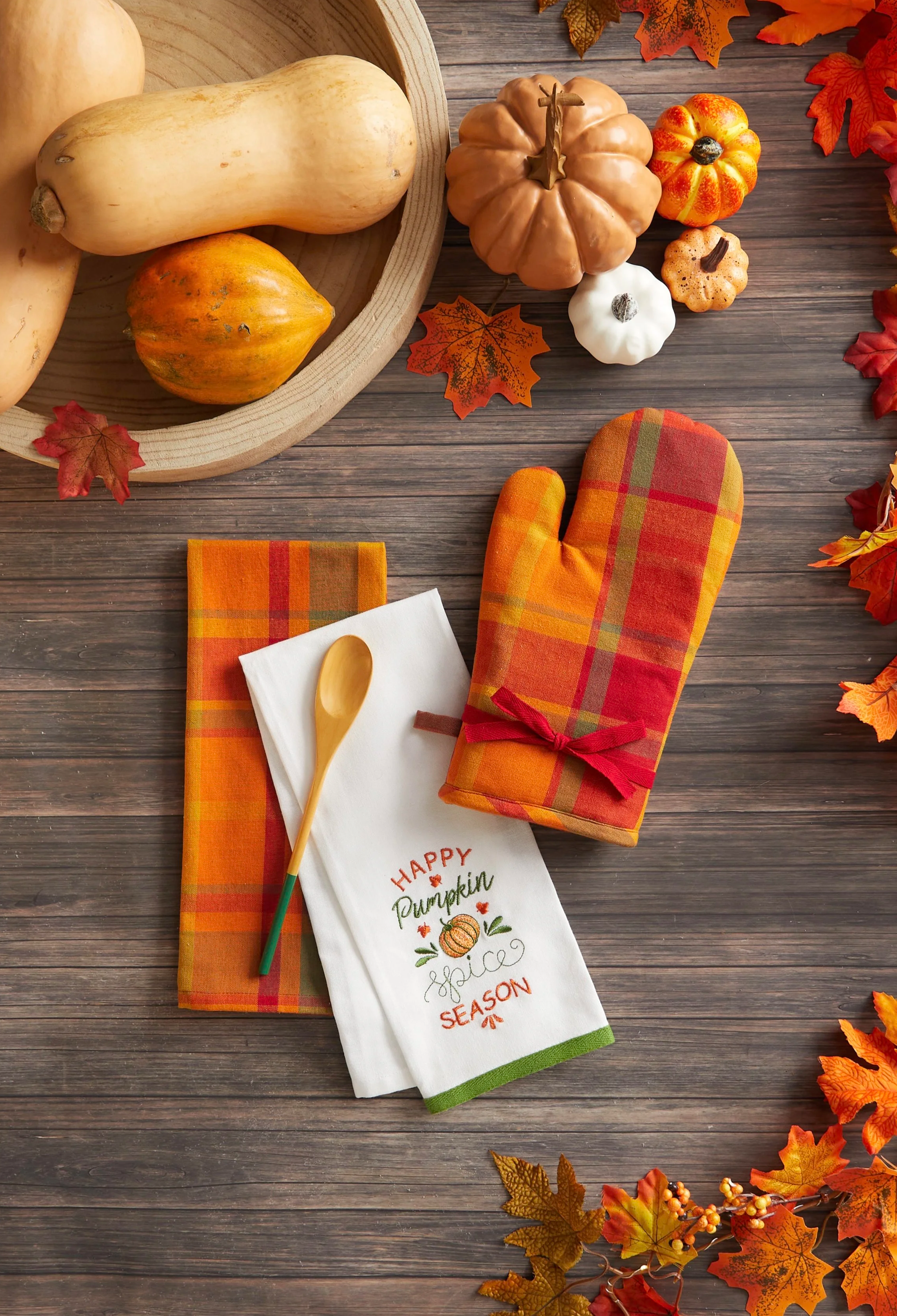 A wooden table decorated with fall-themed items including a paper towel with pumpkin design, orange and yellow fall leaves, a white mitten with orange, green, and red accents, a plaid oven mitt, a white napkin, a wooden spoon, and several small decor