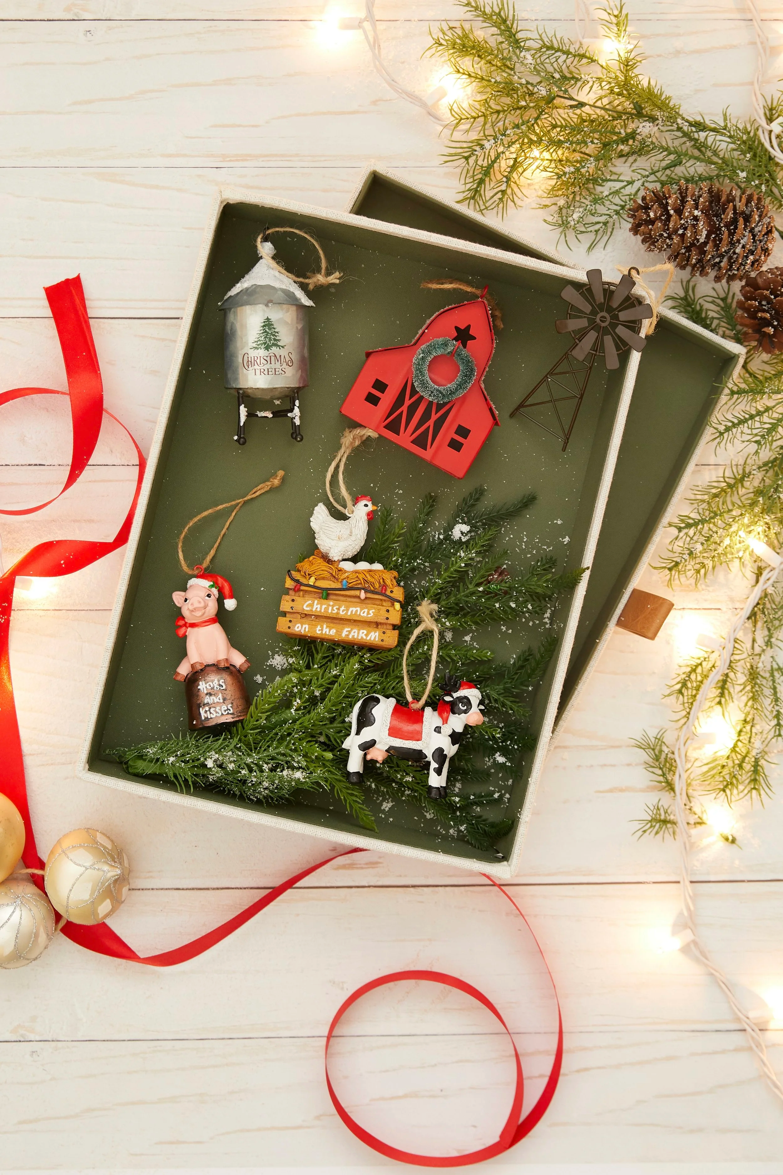 A box of Christmas ornaments and decorations on a white wooden surface decorated with pine branches, pinecones, red ribbon, and string lights.