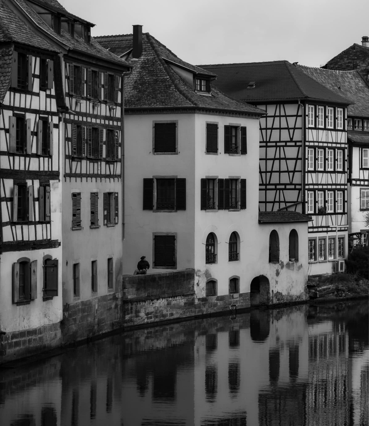 Black and white photo of old European-style buildings along a canal, with a person sitting on the canal wall.