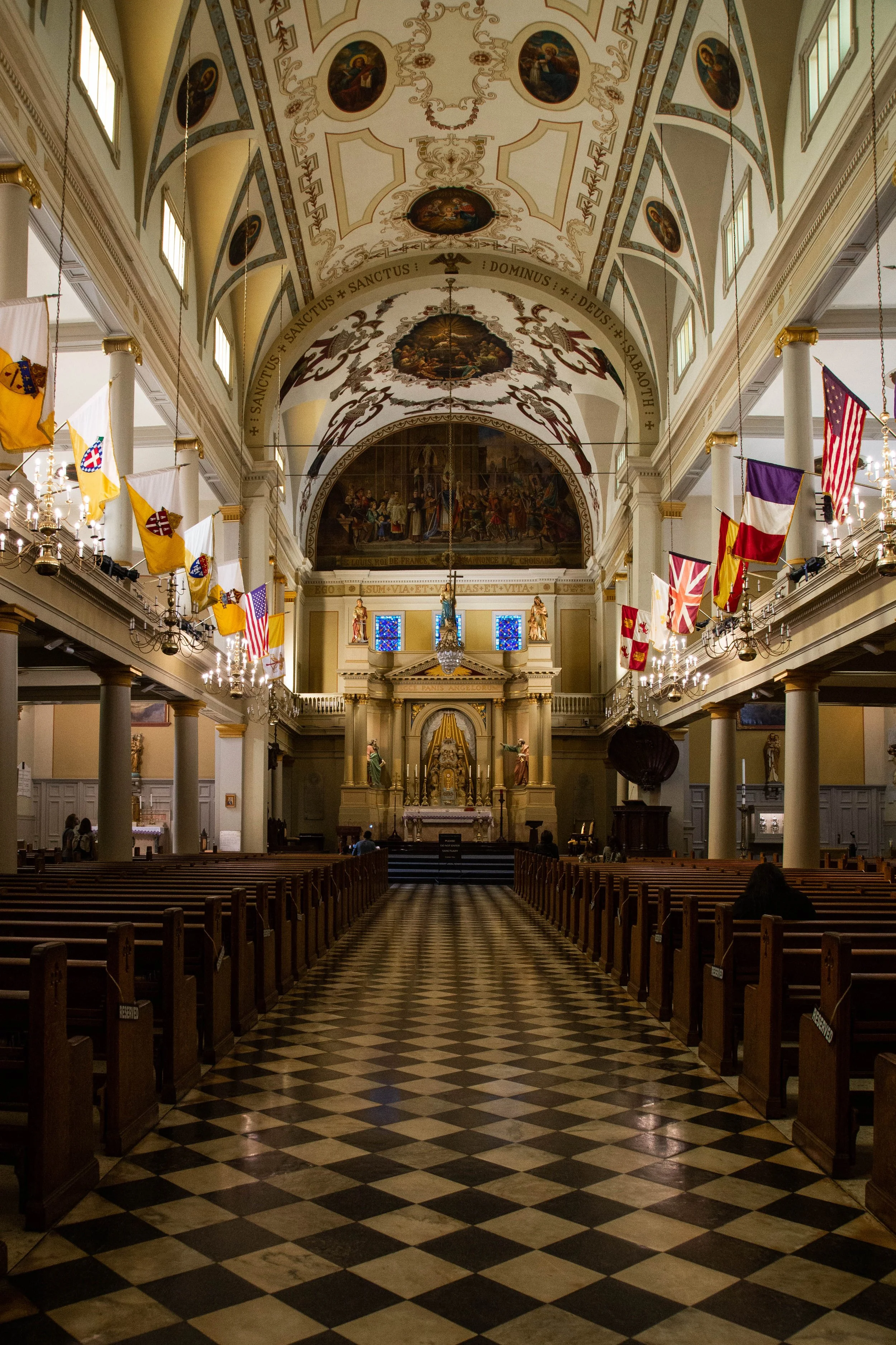 Interior of a church with rows of wooden pews, a checkered floor, and a decorated altar with statues. The ceiling has ornate paintings and religious inscriptions, with flags hanging along the sides.