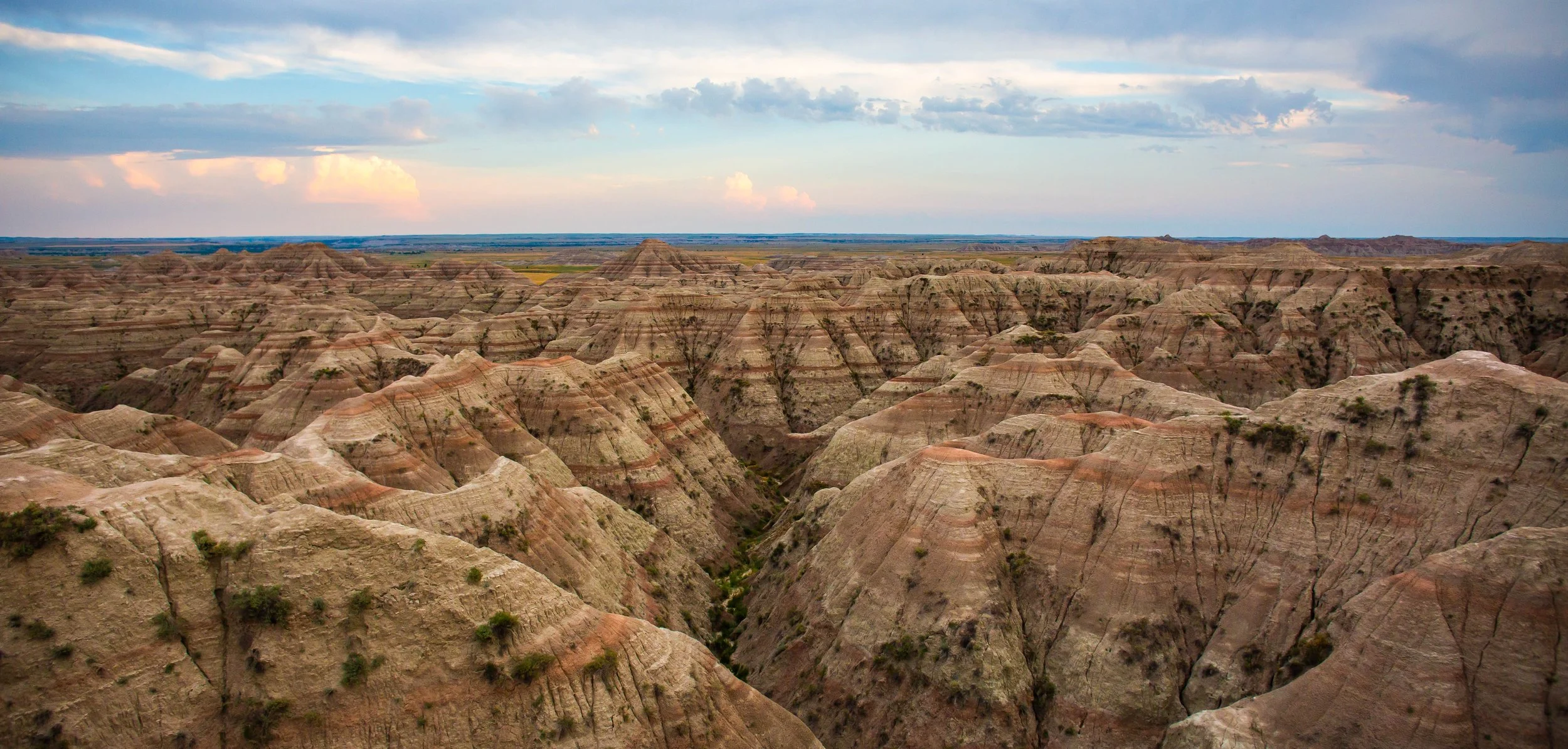 A vast landscape of eroded buttes, hills, and canyons with multicolored layers, under a partly cloudy sky.