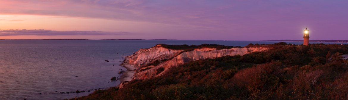 Sunset view of a lighthouse on a rocky coast with pink cliffs and a body of water in the background.