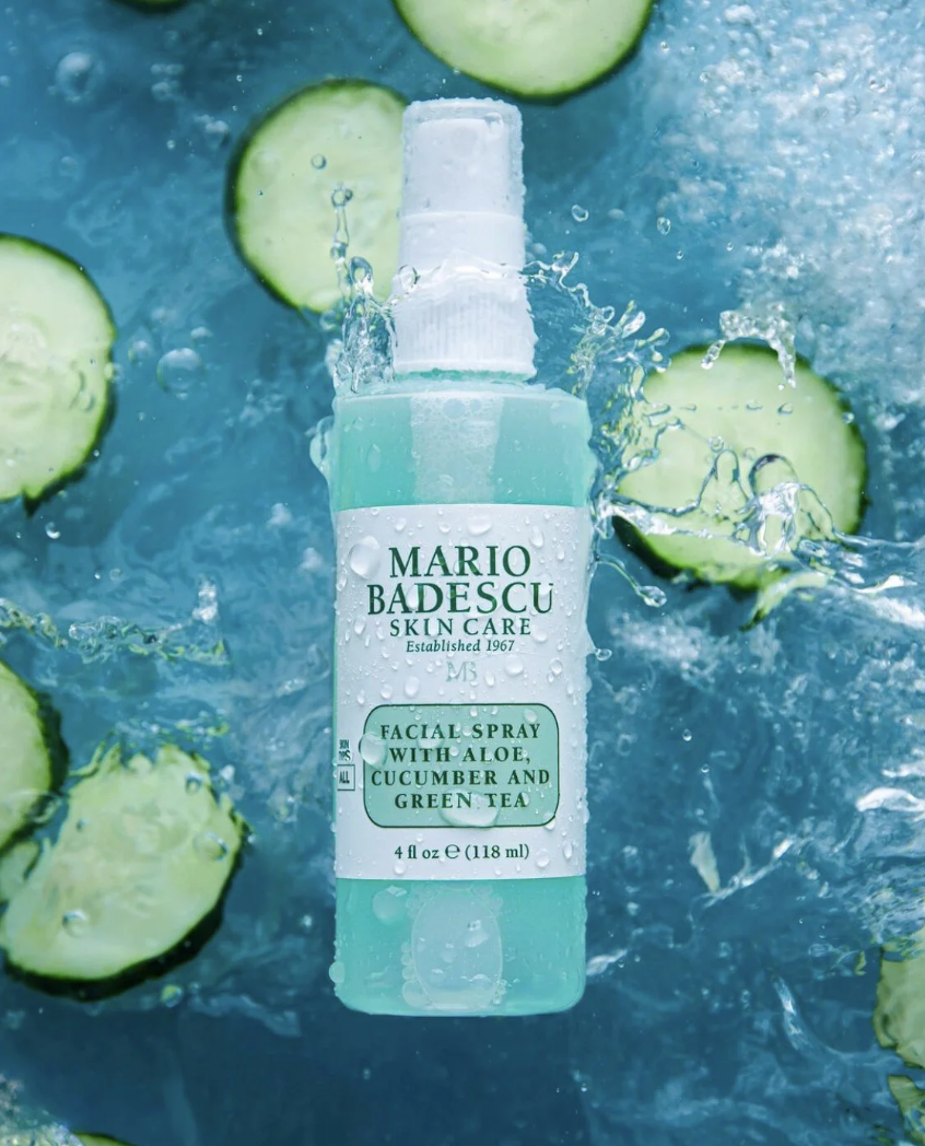 A spray bottle labeled 'Mario Badescu Skin Care Facial Spray with Aloe, Cucumber and Green Tea' surrounded by cucumber slices and splashing water.